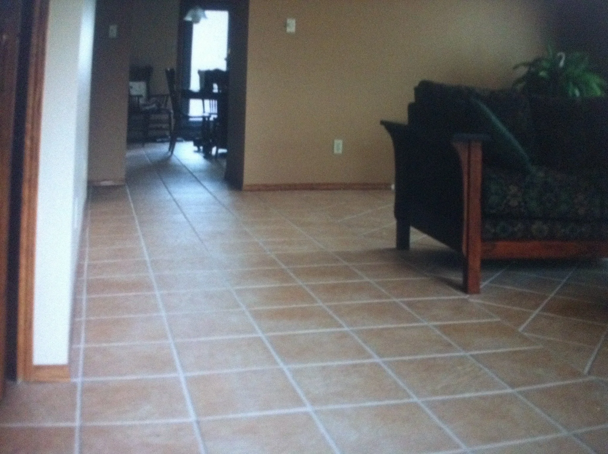 View of a living room with tiled floor, a black sofa with wooden arms and a green cushion, a wall with a light switch, and an open doorway leading to a dining area with a dark wooden table and chairs near a window.