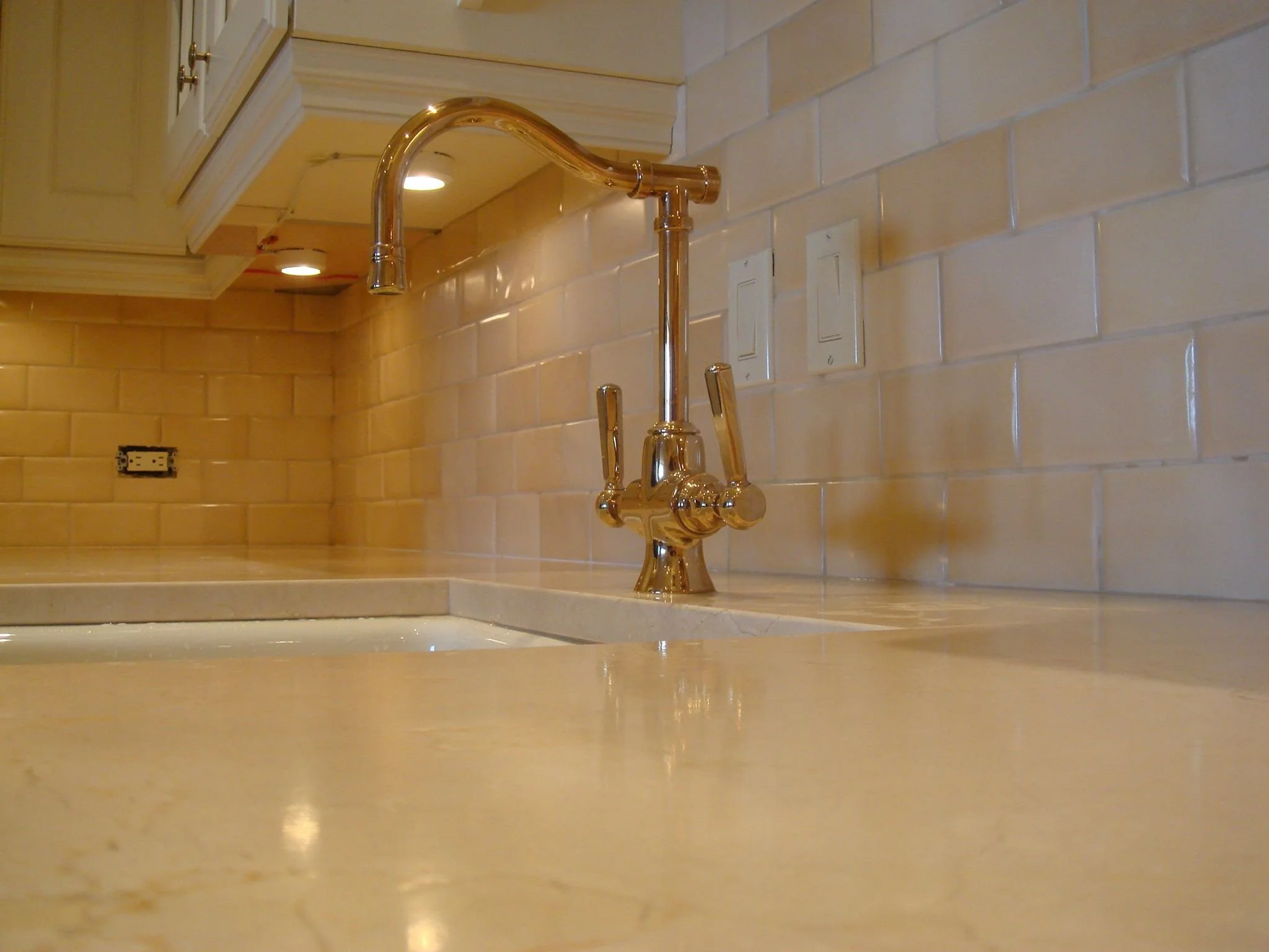 Close-up of a kitchen countertop with a gold-tone faucet, cream-colored tiled backsplash, and wooden cabinets above.