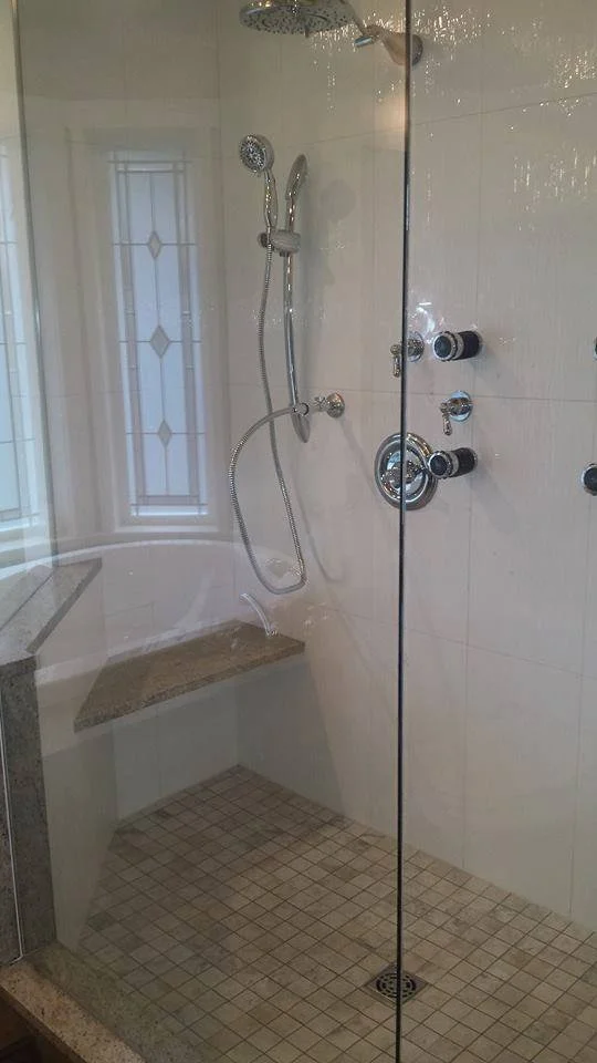 A walk-in bathroom shower with a granite bench, beige tiled walls, and a glass door. The shower has a handheld showerhead, a fixed showerhead, and multiple black and chrome control knobs.