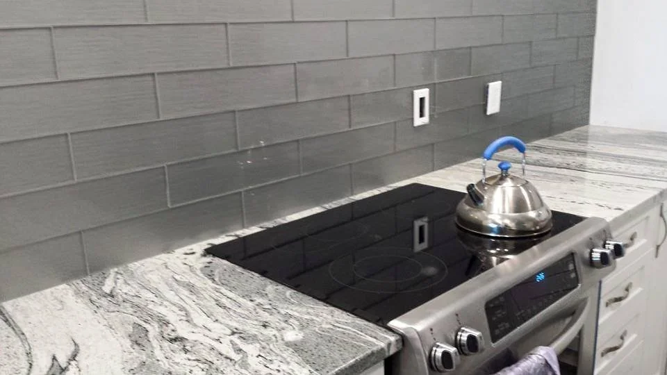 Kitchen stove with a stainless steel kettle on top, gray tile backsplash, and granite countertops.