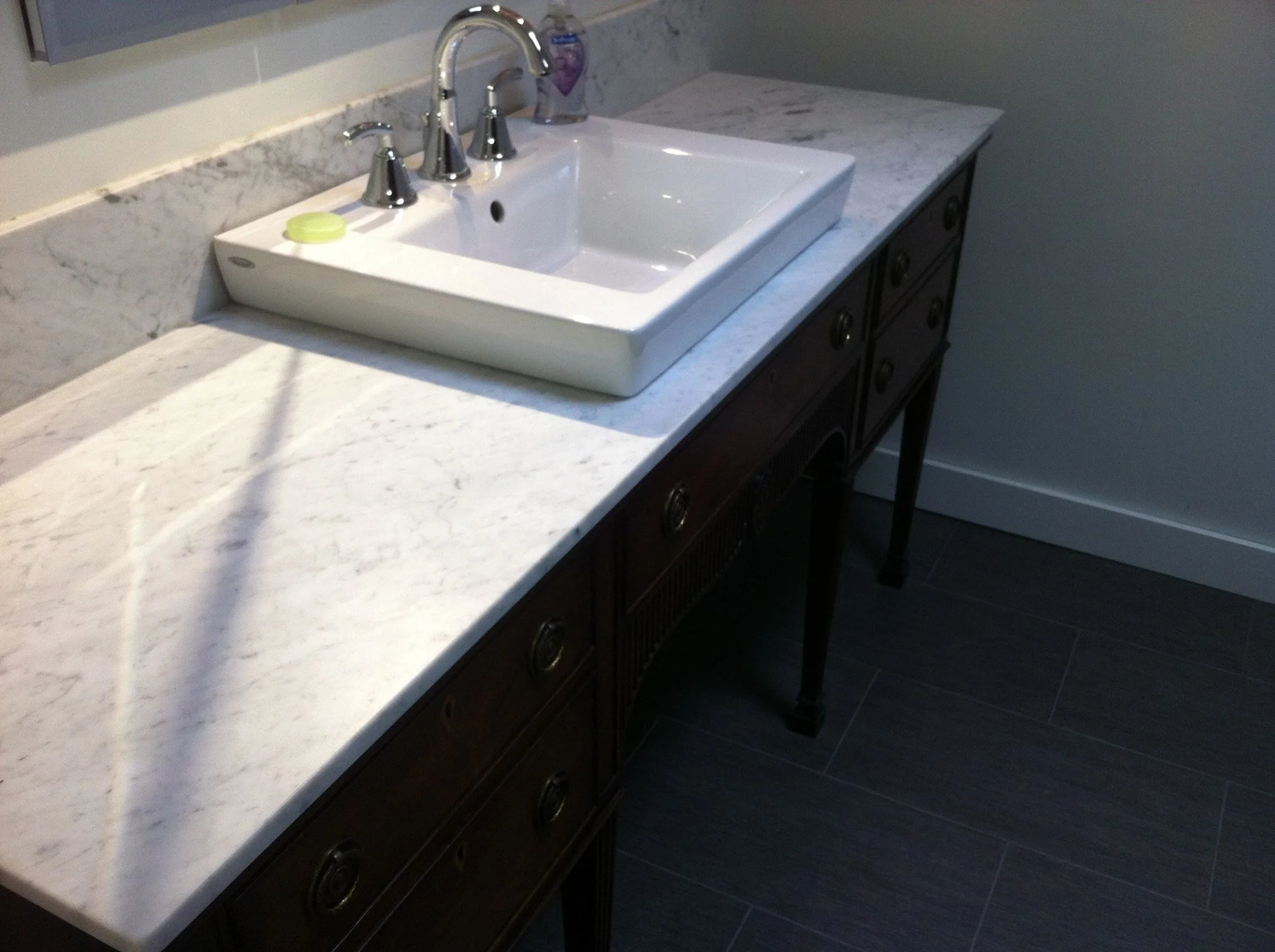 Bathroom vanity with a white marble countertop, a rectangular vessel sink, and a three-piece chrome faucet. Dark wooden cabinet with multiple drawers and black legs, with a soap dispenser and soap dish on the countertop.