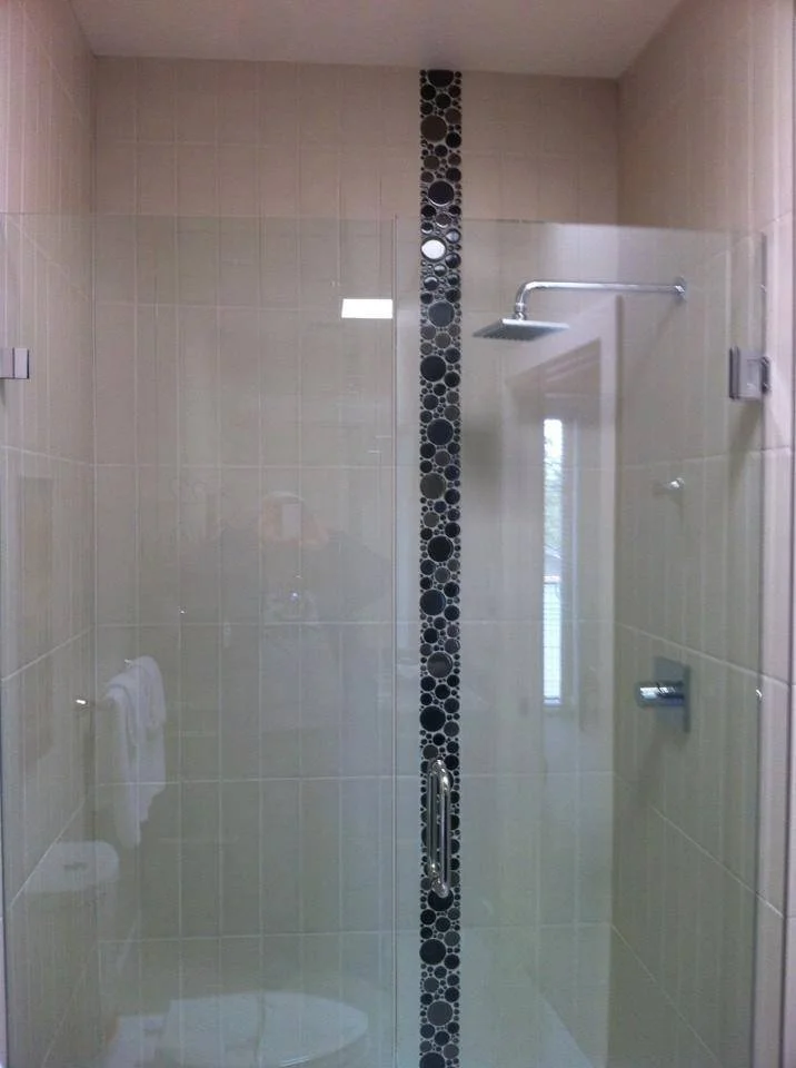 Clean glass shower door with beige tiled walls, a black decorative vertical strip, a silver shower head, and a frosted glass window reflected on the glass.
