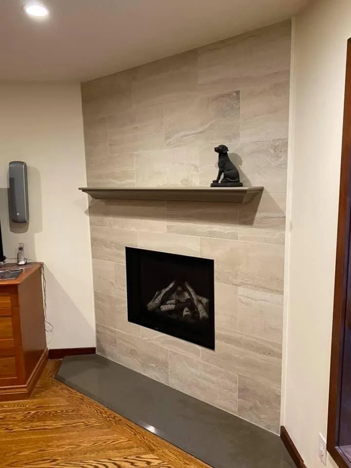 A modern fireplace with a beige tiled wall and a shelf holding a small dog figurine. The fireplace is framed in black and set into the tiled wall, with a gray hearth below.