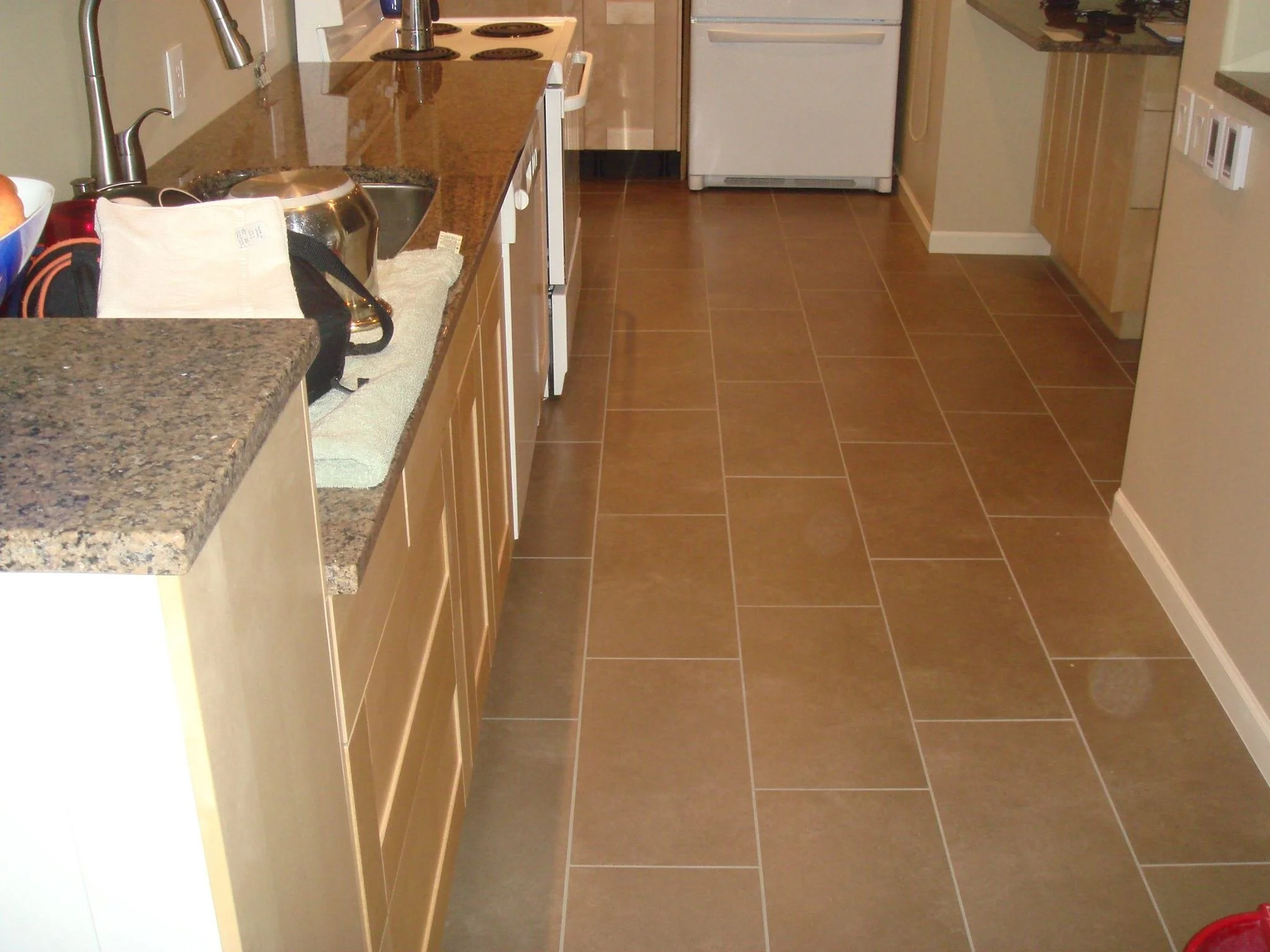 A kitchen with brown tiled flooring, wooden cabinetry, a granite countertop, a white refrigerator, and various small kitchen items on the counter.