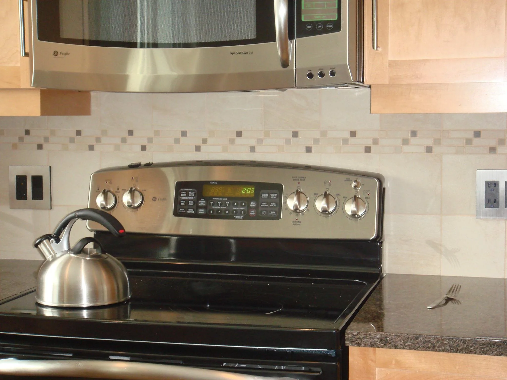 Stainless steel electric stove with a black cooktop, a tea kettle on the left burner, and four control knobs. Microwave oven mounted above with digital display, situated in a kitchen with beige tiled backsplash and wooden cabinets.