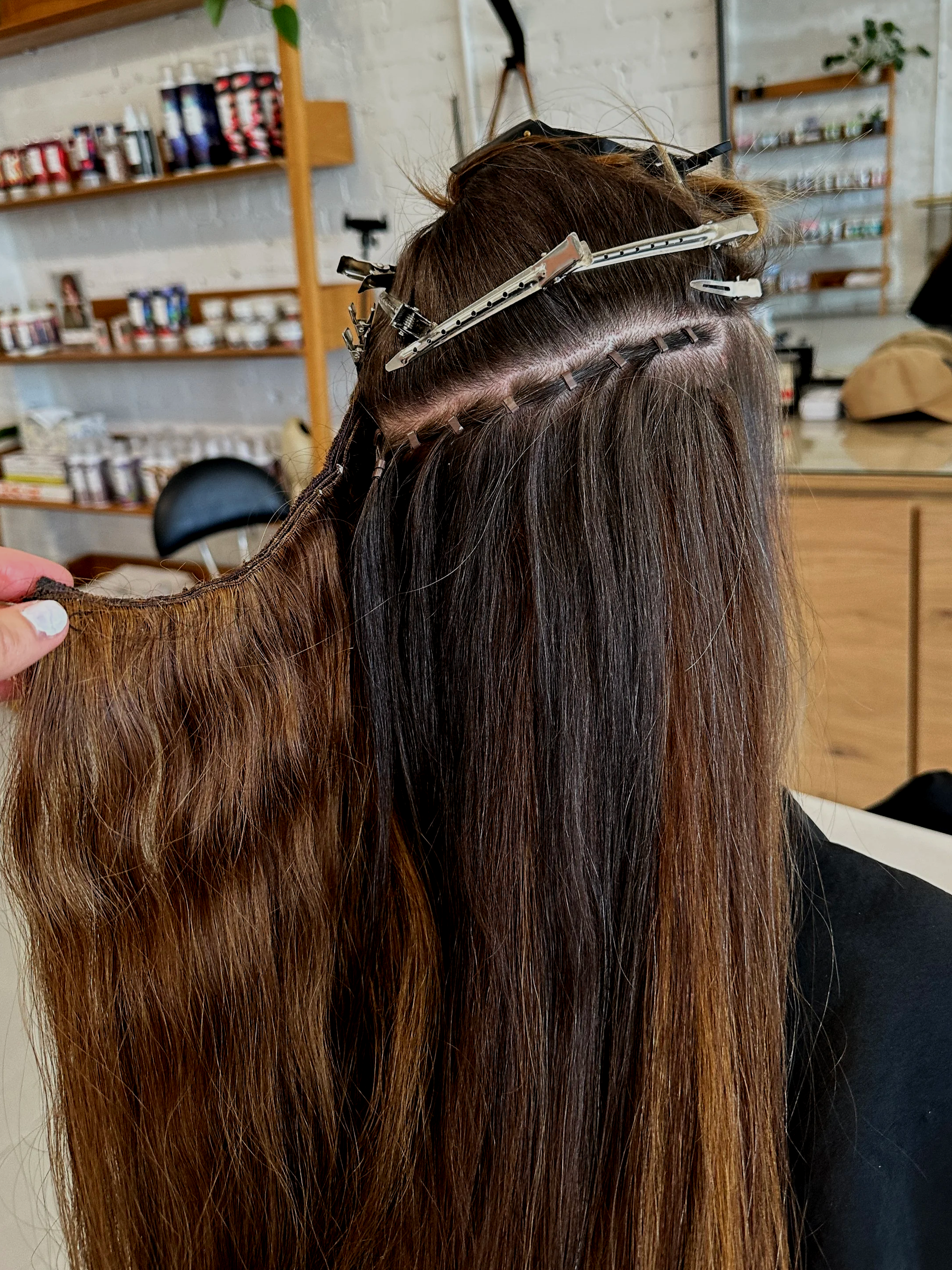 Back view of a woman with long brown hair in the process of getting a hairstyle done, with hair clips securing sections of her hair.