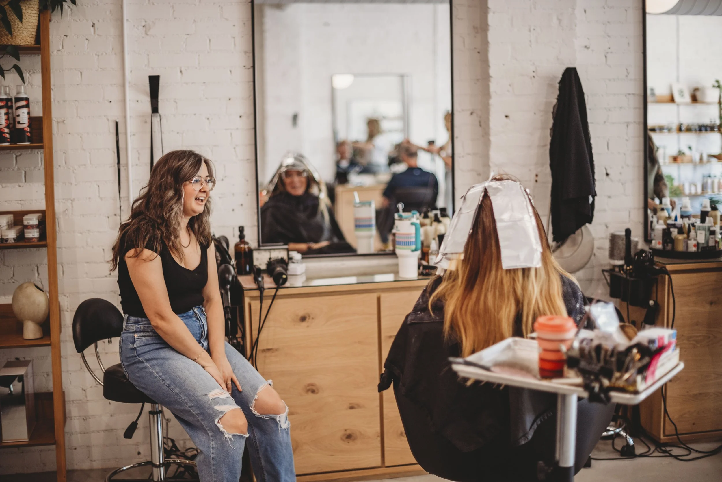 A woman with glasses and long wavy hair sitting on a white stool, wearing a floral top and black pants, smiling. In the background, another woman with blonde hair talking and gesturing, sitting at a white dresser with a mirror, lamp, and bottles.