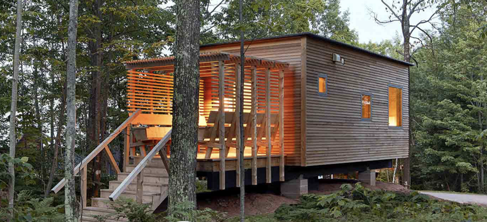 A modern wooden house elevated on stilts, with a front staircase, surrounded by trees and greenery, with warm interior lighting visible through the windows. The nest lodging option at our reading retreats.