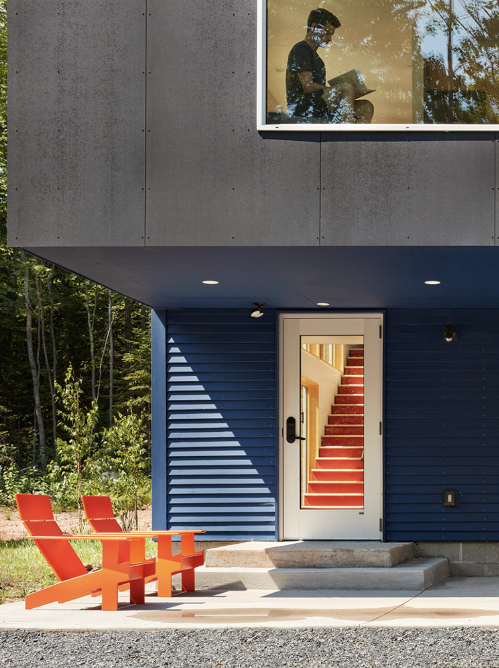 Modern house at reading retreat with gray upper siding and blue lower siding, glass door with interior staircase visible through it, red outdoor stairs, red chairs outside, surrounded by trees.