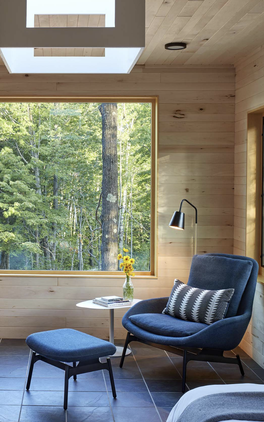A cozy reading nook with a blue armchair and matching ottoman near a large window overlooking a lush green forest at our reading retreat. Natural light floods the space through the window and skylight.
