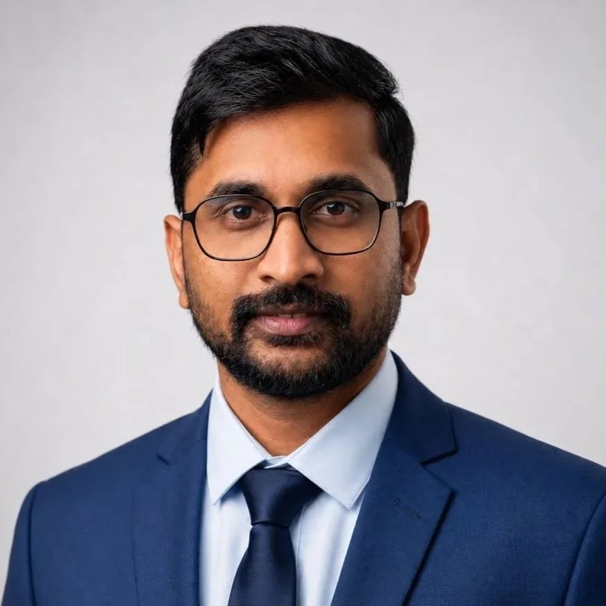 Professional headshot of a man in a blue suit, white shirt, and dark tie, wearing glasses, against a plain background.