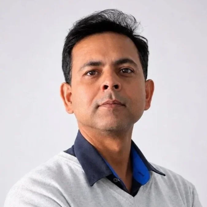 A man with short black hair and a slight smile, wearing a gray sweater over a dark blue collared shirt, stands against a plain light gray background.