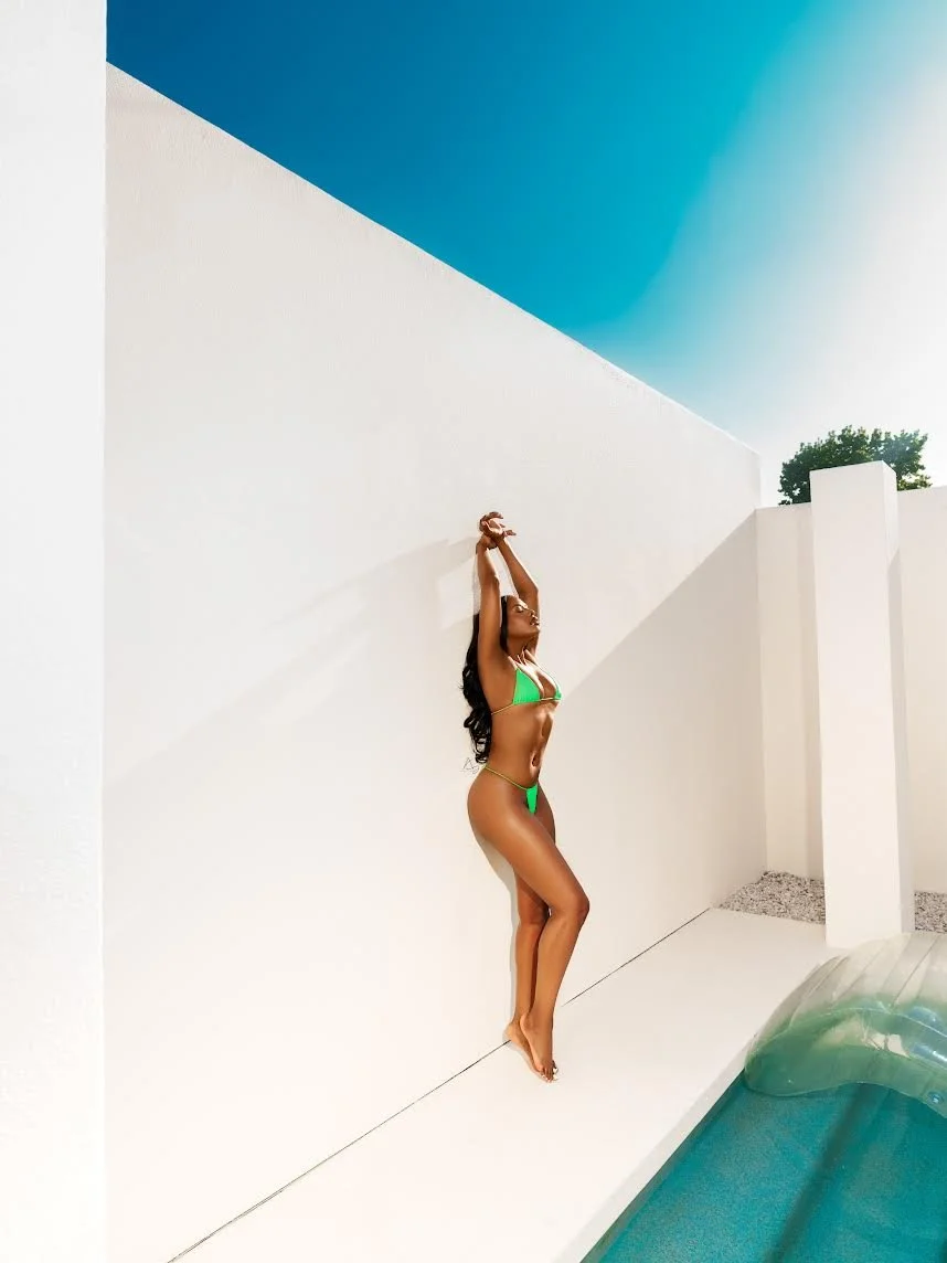 A woman in a green bikini standing on the edge of a pool area with her arms raised above her head, leaning against a white wall under a clear blue sky.