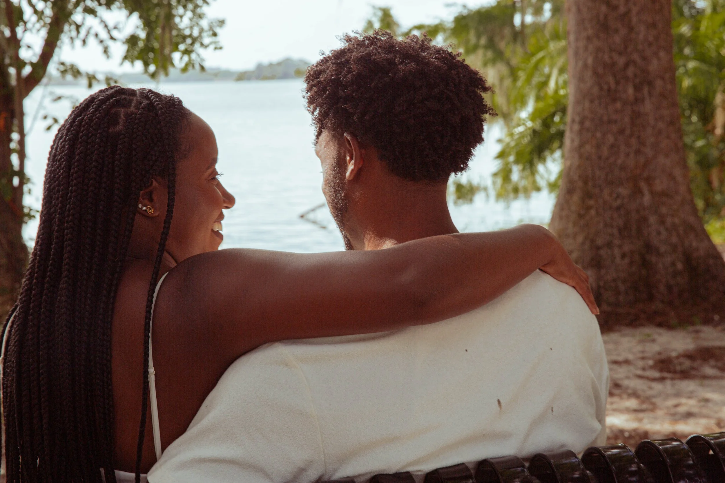 Couple with one person having their arm around another looking at each other seated in front a body of water 