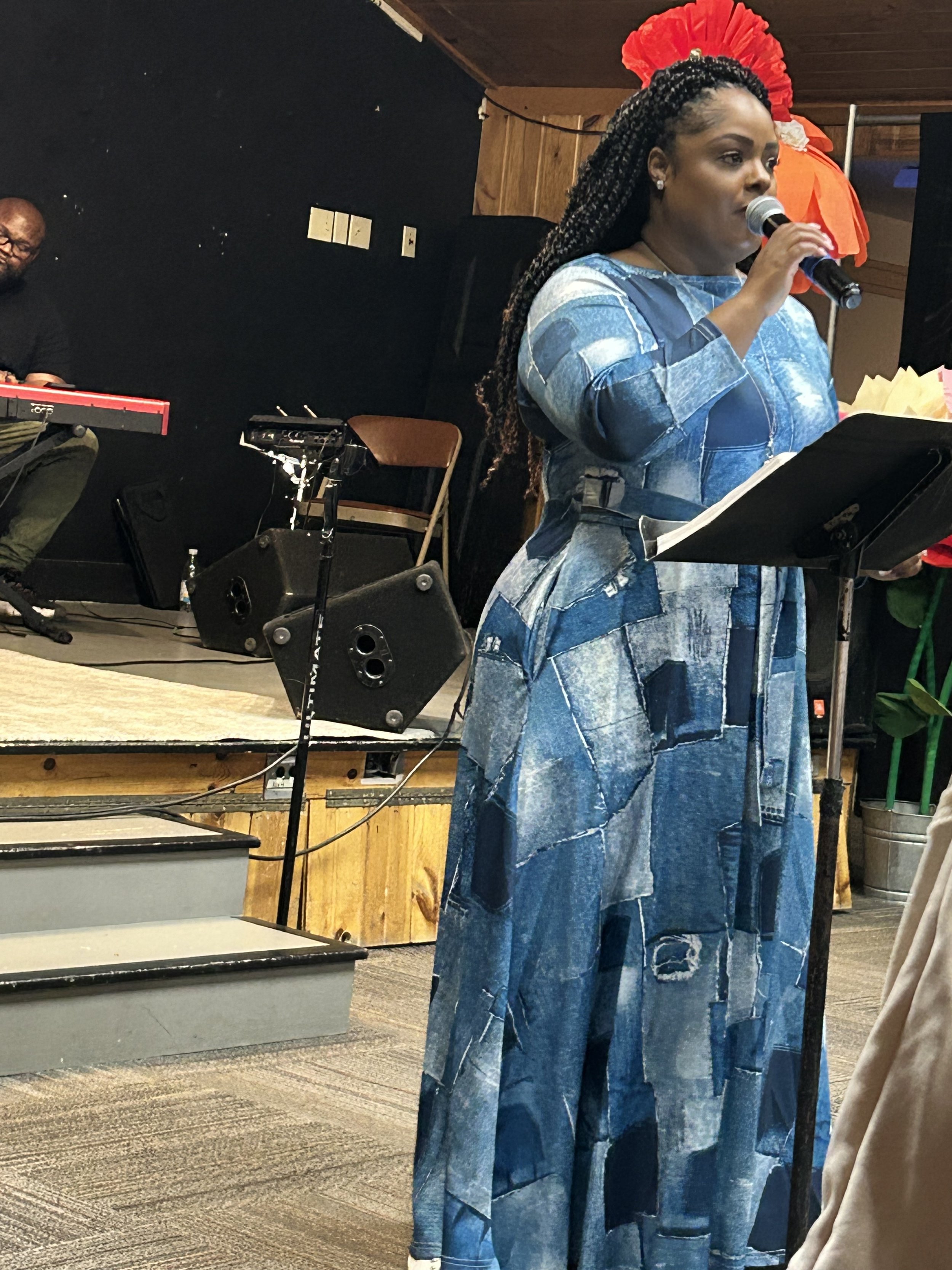 A woman with braided hair is singing into a microphone while holding sheet music, wearing a blue patchwork dress. A man playing a keyboard is visible in the background, along with stage equipment and a wooden wall.