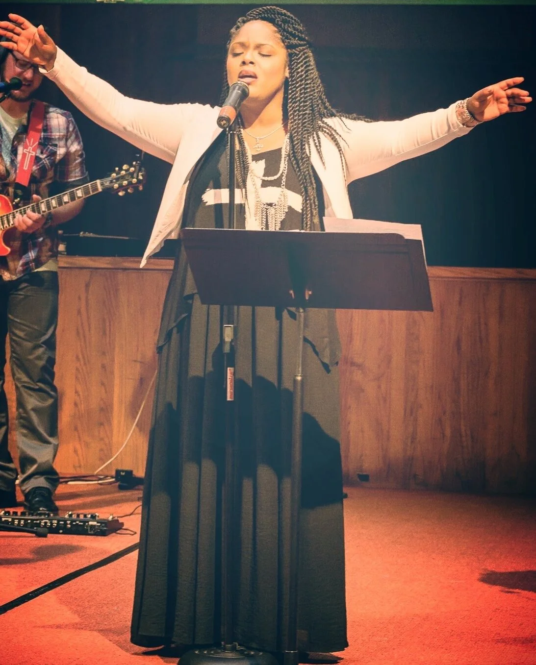 A woman singing with her arms outstretched, standing at a music stand, with a musician playing guitar beside her on a stage with wooden paneling.