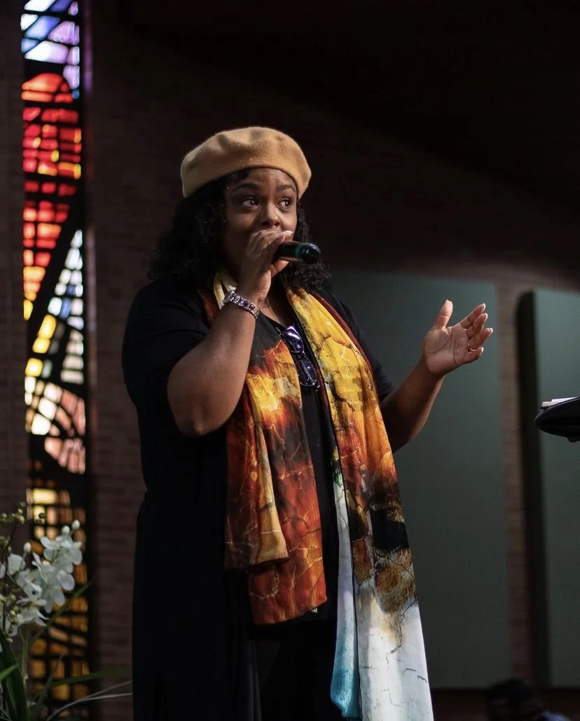 A woman with dark curly hair wearing a beige beret and colorful scarf, speaking into a microphone inside a building with stained glass windows.