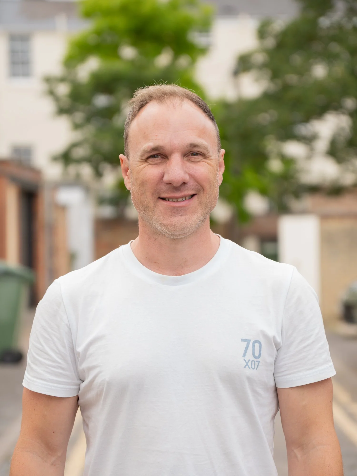 A smiling man with short hair and light skin, wearing a white T-shirt, standing outdoors with green trees and residential buildings in the background.