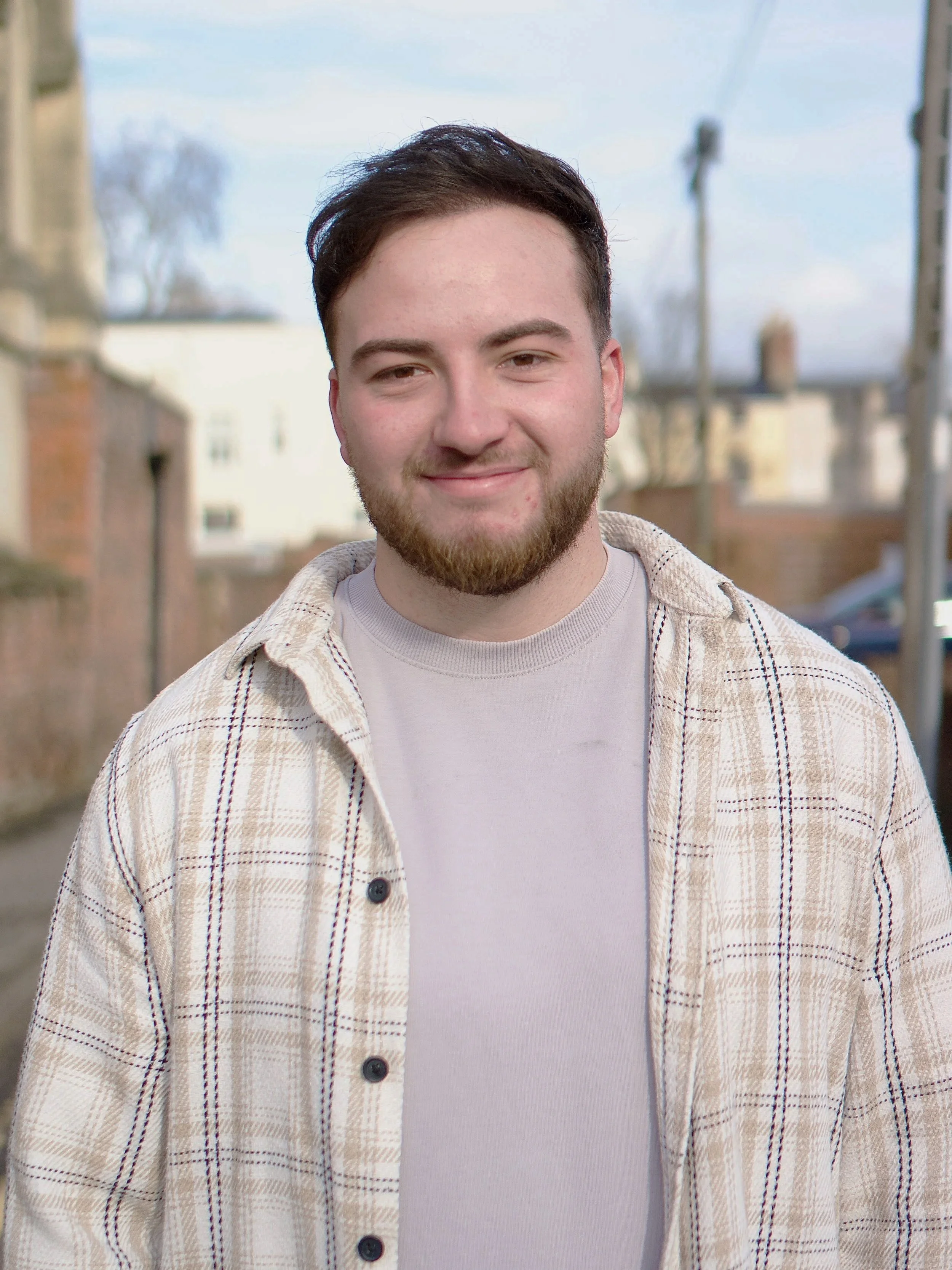 A young man with short dark hair and a beard standing outdoors in residential area, wearing a beige plaid shirt over a light gray T-shirt, smiling at the camera.