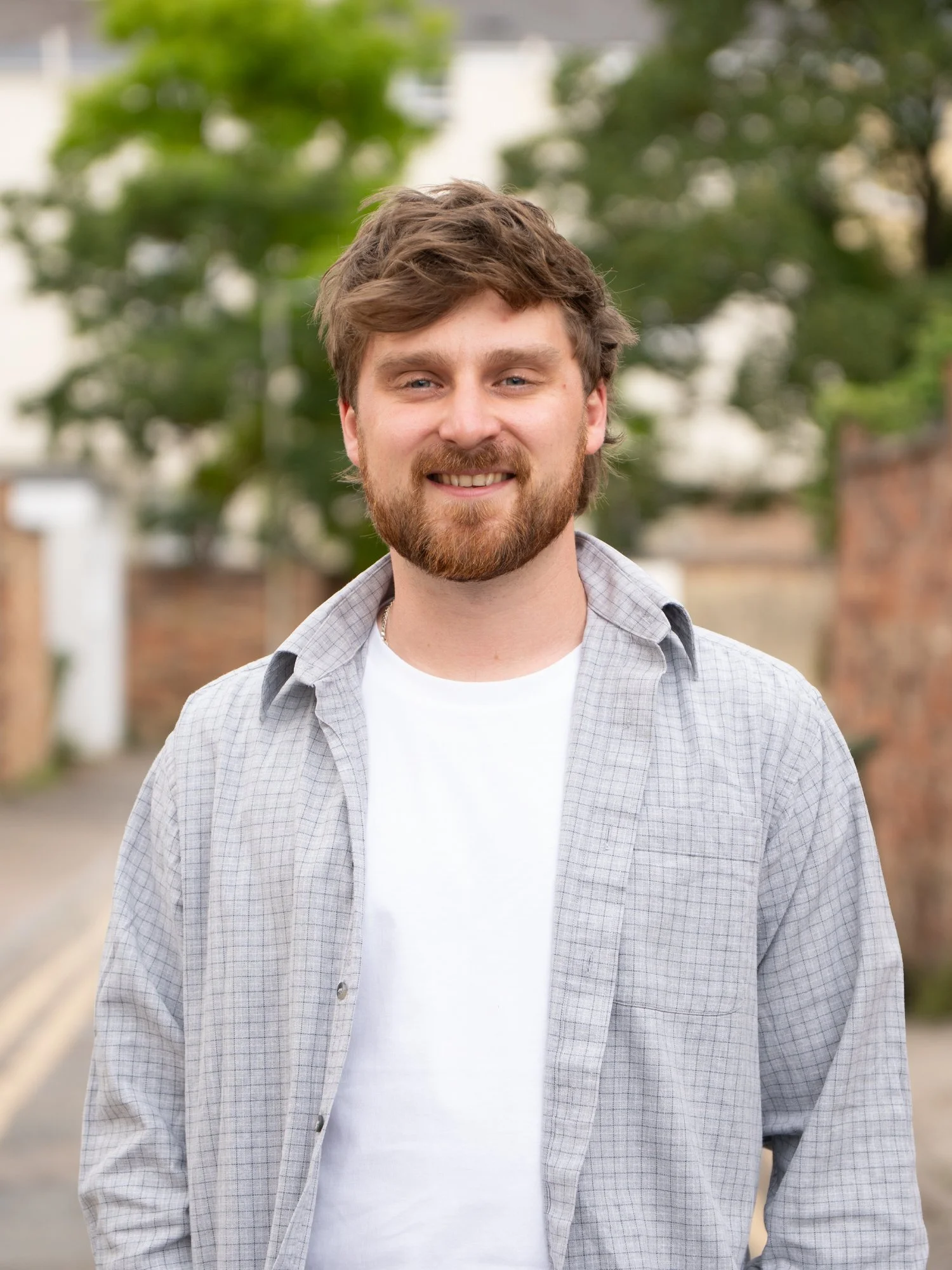 A young man with brown hair and a beard, smiling, standing outdoors with blurred trees and buildings in the background.