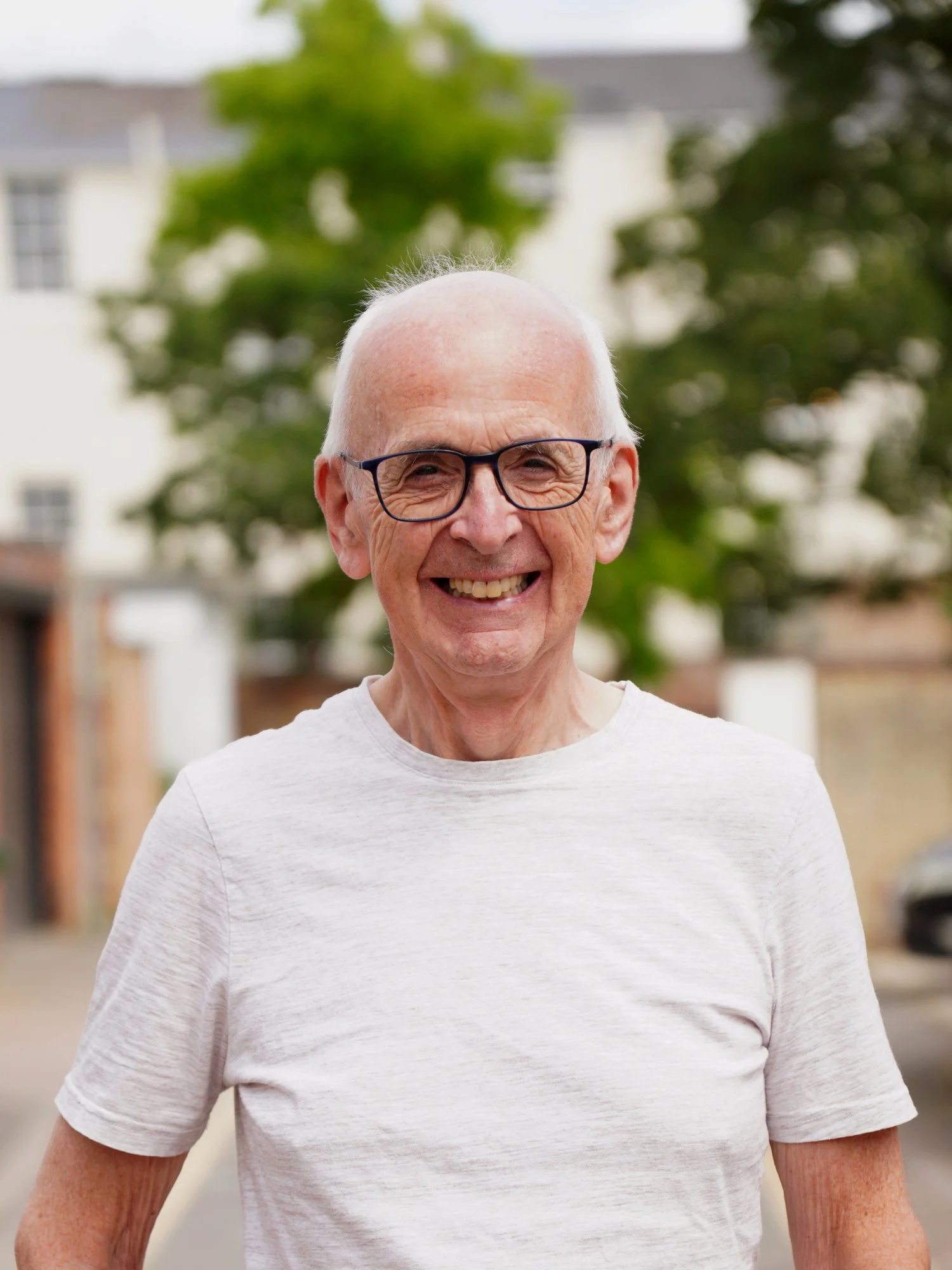 Close-up of an elderly man with white hair and glasses smiling outdoors, wearing a light gray t-shirt, with trees and a building in the background.