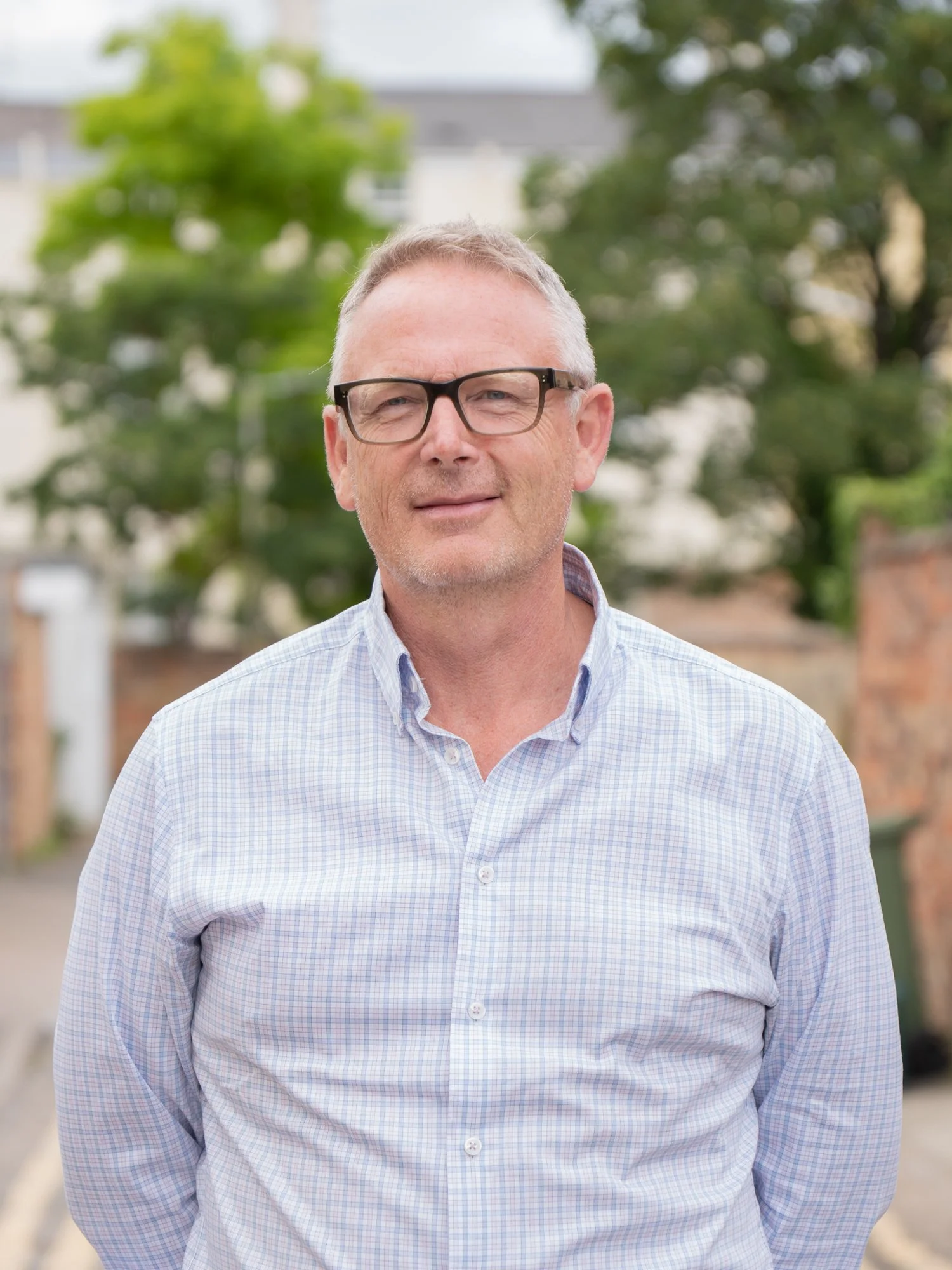 A middle-aged man with short gray hair, black glasses, and a light blue checkered shirt standing outdoors with trees and buildings in the background.