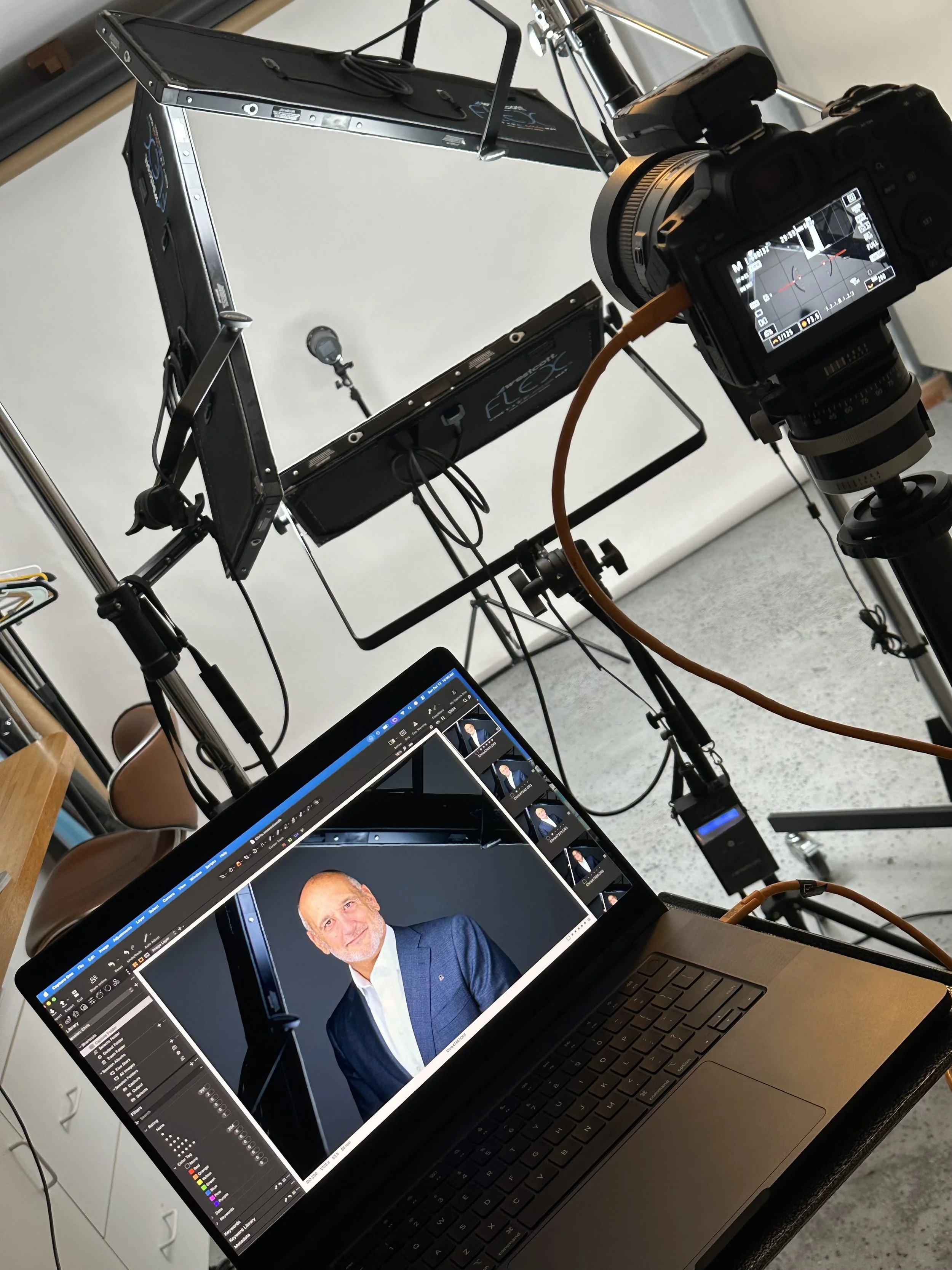 Photography setup with a camera on a tripod, an open laptop displaying a professional portrait, and a lighting reflector, indicating a photo shoot environment.