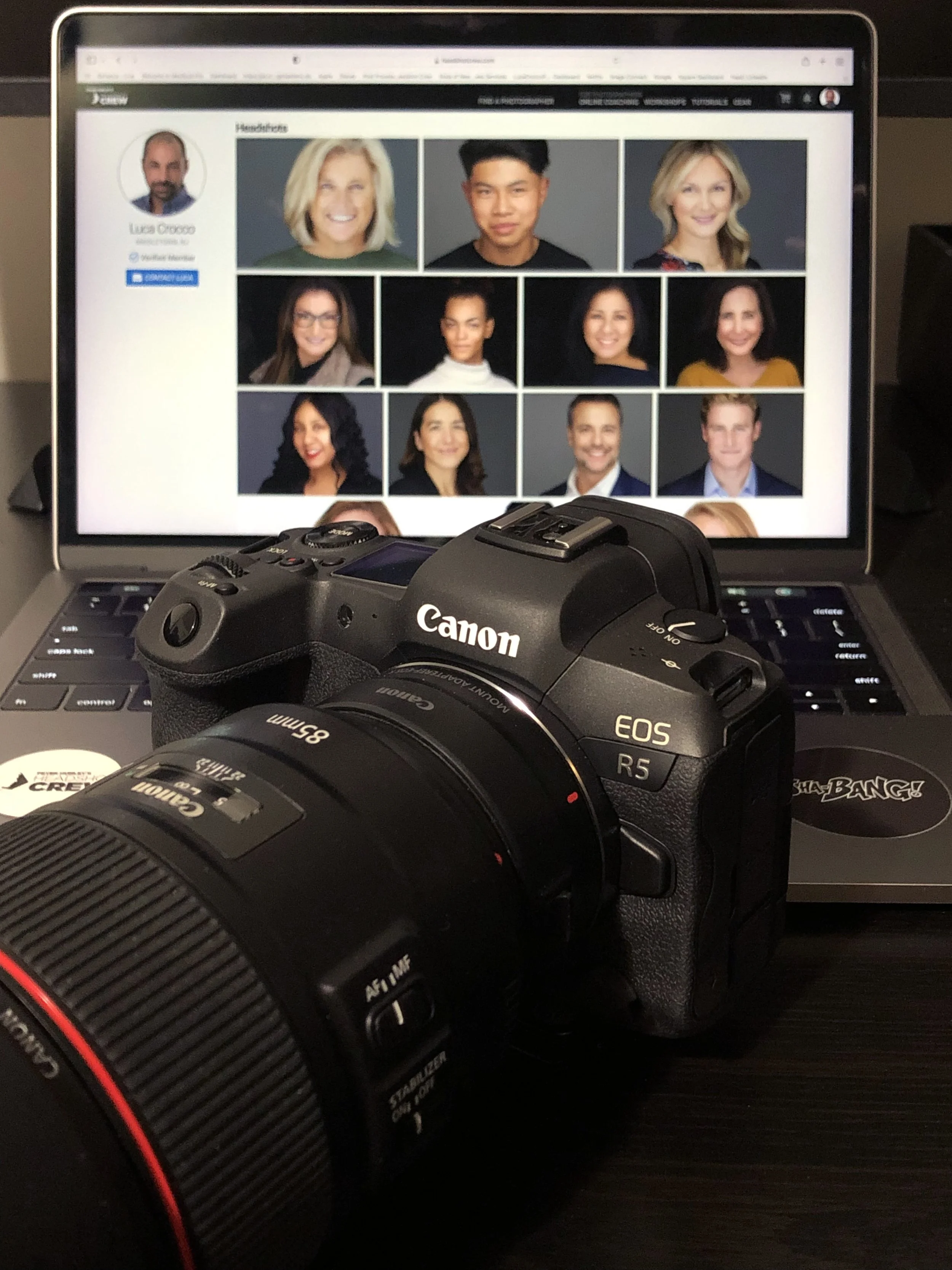A DSLR camera placed on a laptop keyboard, with the laptop screen displaying a webpage with headshots of nine diverse people arranged in a grid.