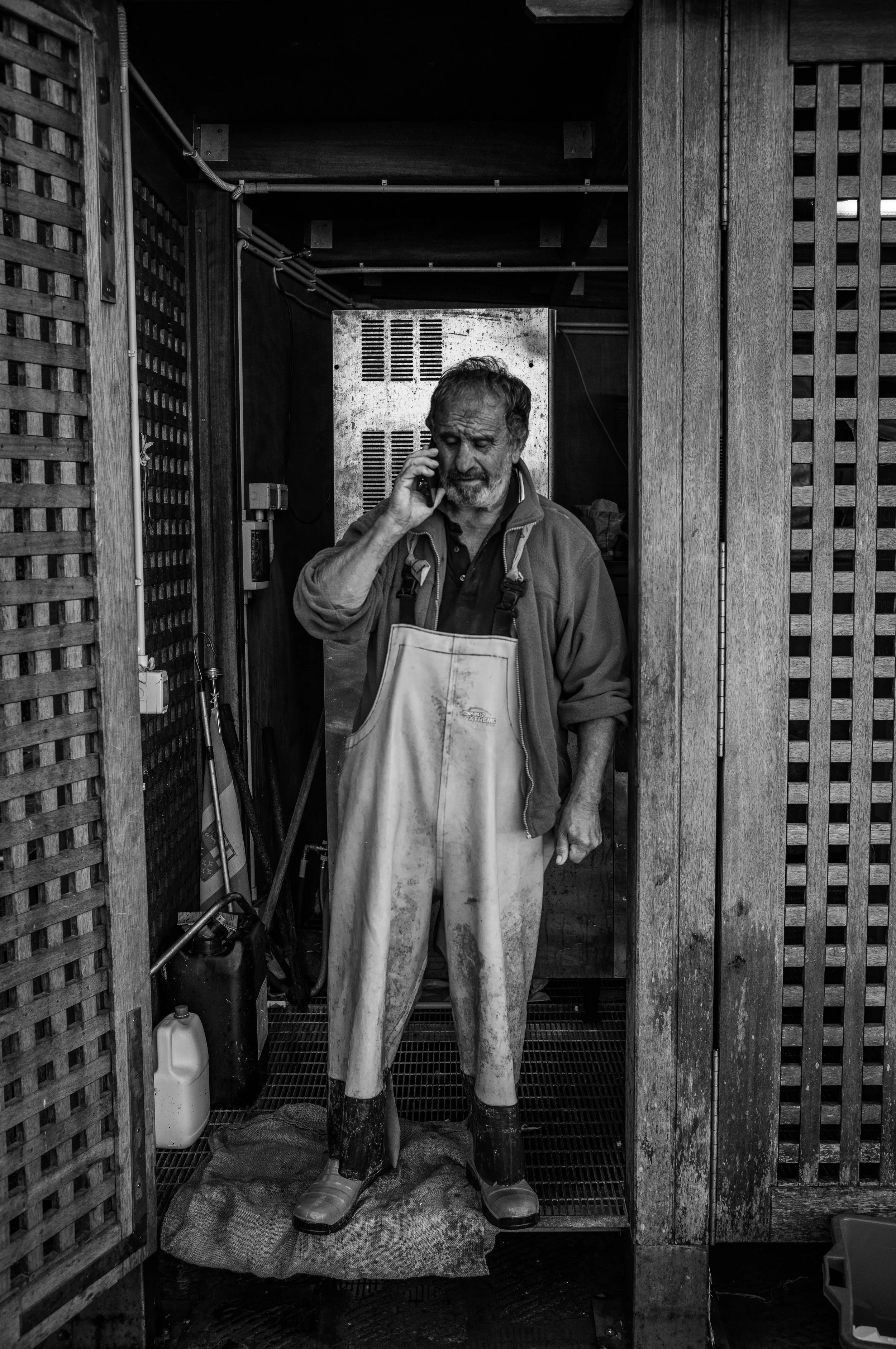 Ein älterer Mann trägt Gummistiefel und Schmutzige Arbeitshose, steht in einem Holzverschlag, spricht am Telefon, im Hintergrund eine metallene Gerätebox.