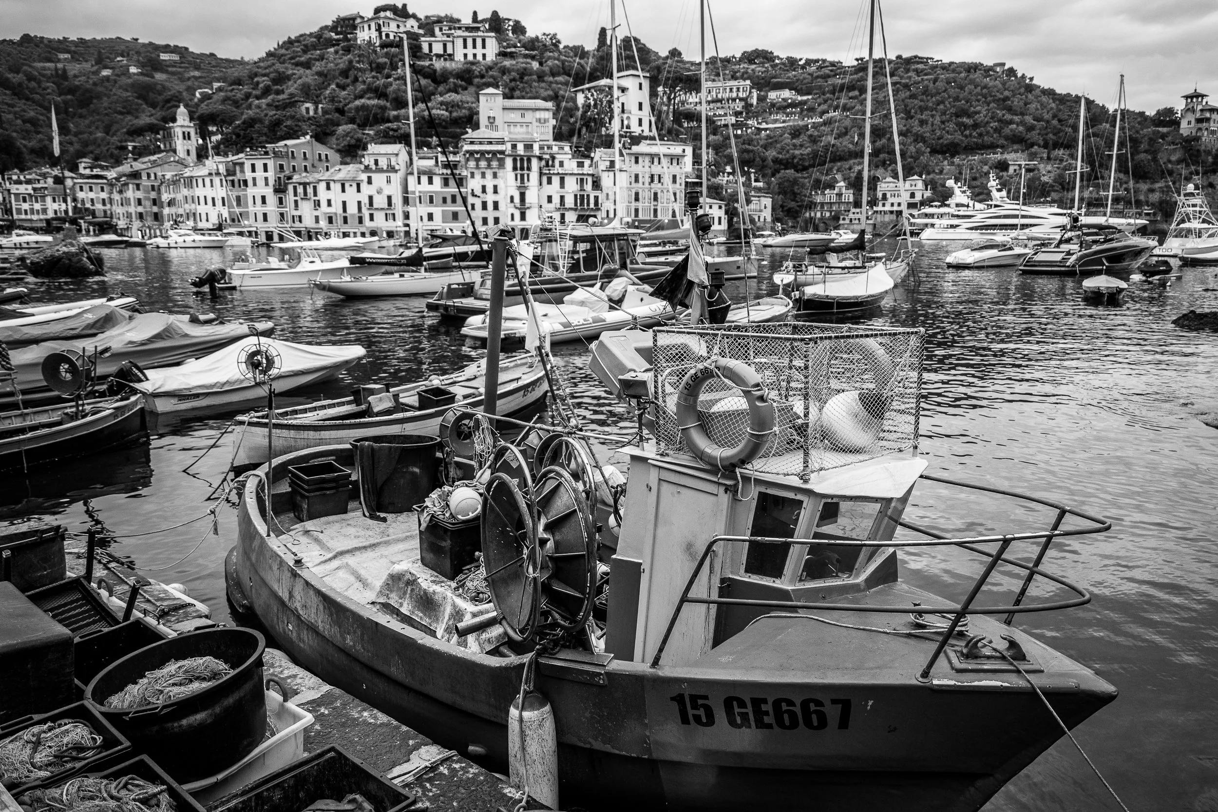 Ein Schwarz-Weiß-Bild eines ruhigen Hafens mit mehreren Booten, einem alten Fischerboot am Kai und einer Stadt im Hintergrund mit Hügeln und Gebäuden.