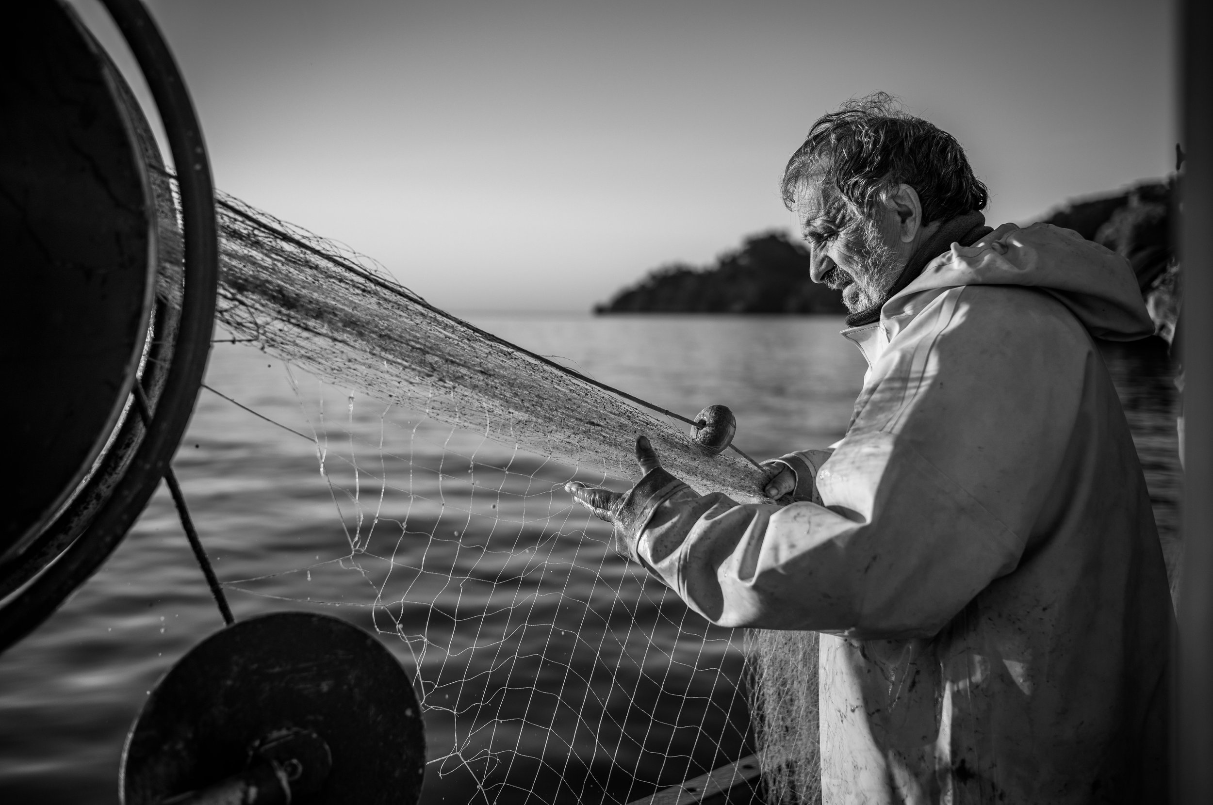 Ein älterer Mann beim Fischfang auf einem Boot, der eine Fischreuse hält, vor einer ruhigen Wasserlandschaft, schwarz-weiß Aufnahme.