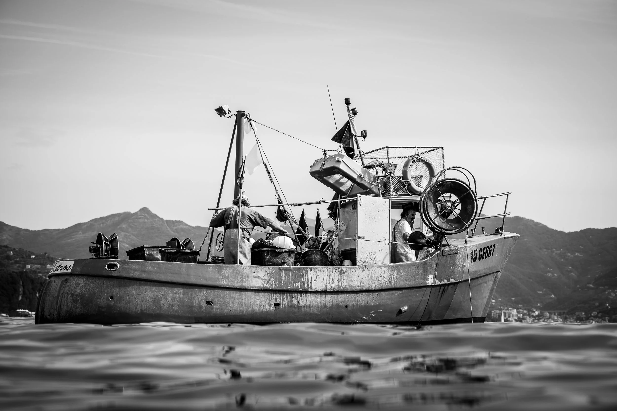 Schwarz-weiß-Foto eines Fischkitts auf dem Wasser mit Bergen im Hintergrund, zwei Männer sind an Bord und arbeiten, es gibt Tauchausrüstung und Rettungsringe auf dem Boot.