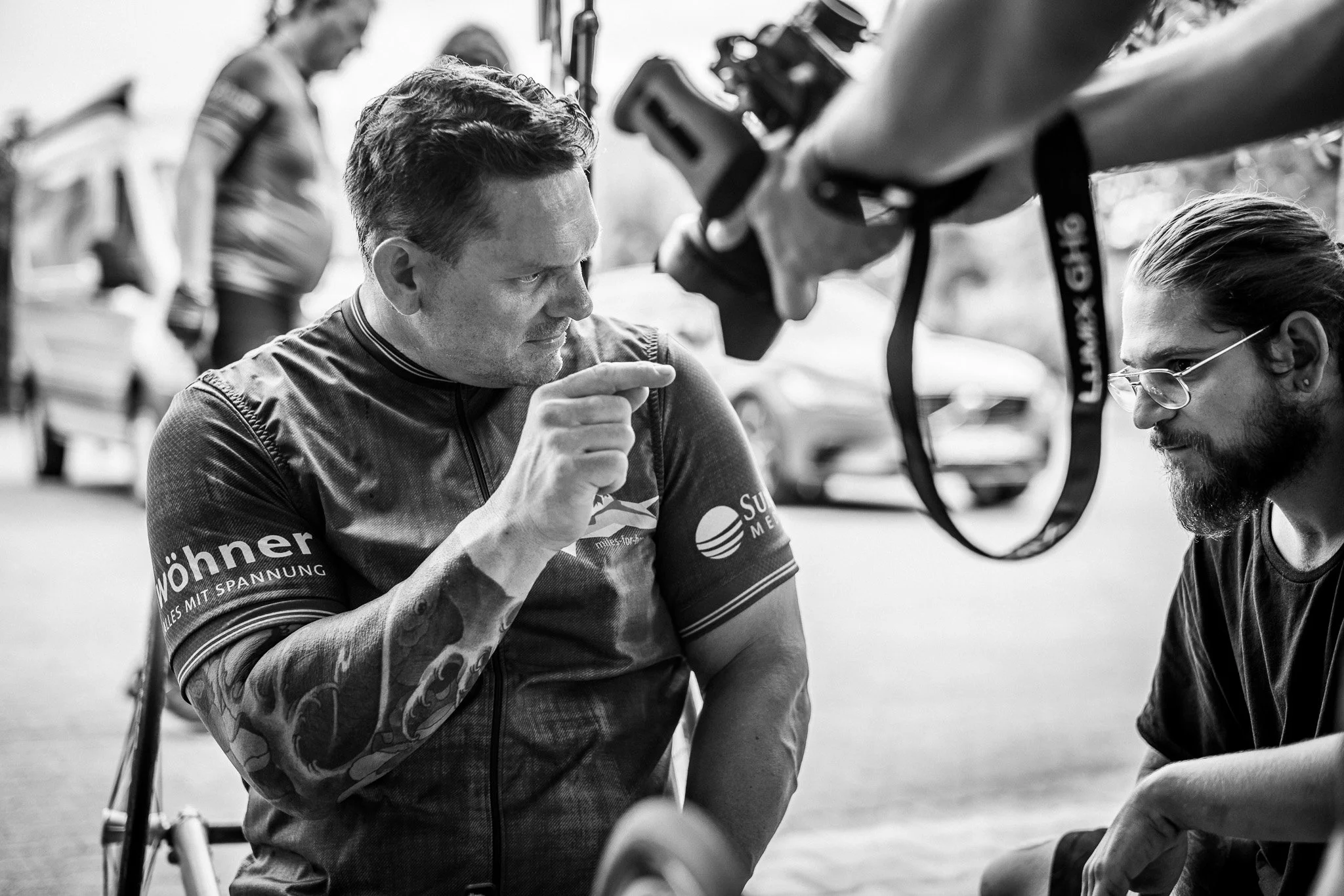 Zwei Männer, einer mit Tattoos auf dem Arm und Rennrad-Kleidung, zeigt auf seinen Arm, während ein Mann mit Brille und dunklem T-Shirt ihm zuhört. Im Hintergrund ist ein Fahrrad und eine Person im Profil. Das Bild ist in Schwarz-Weiß.