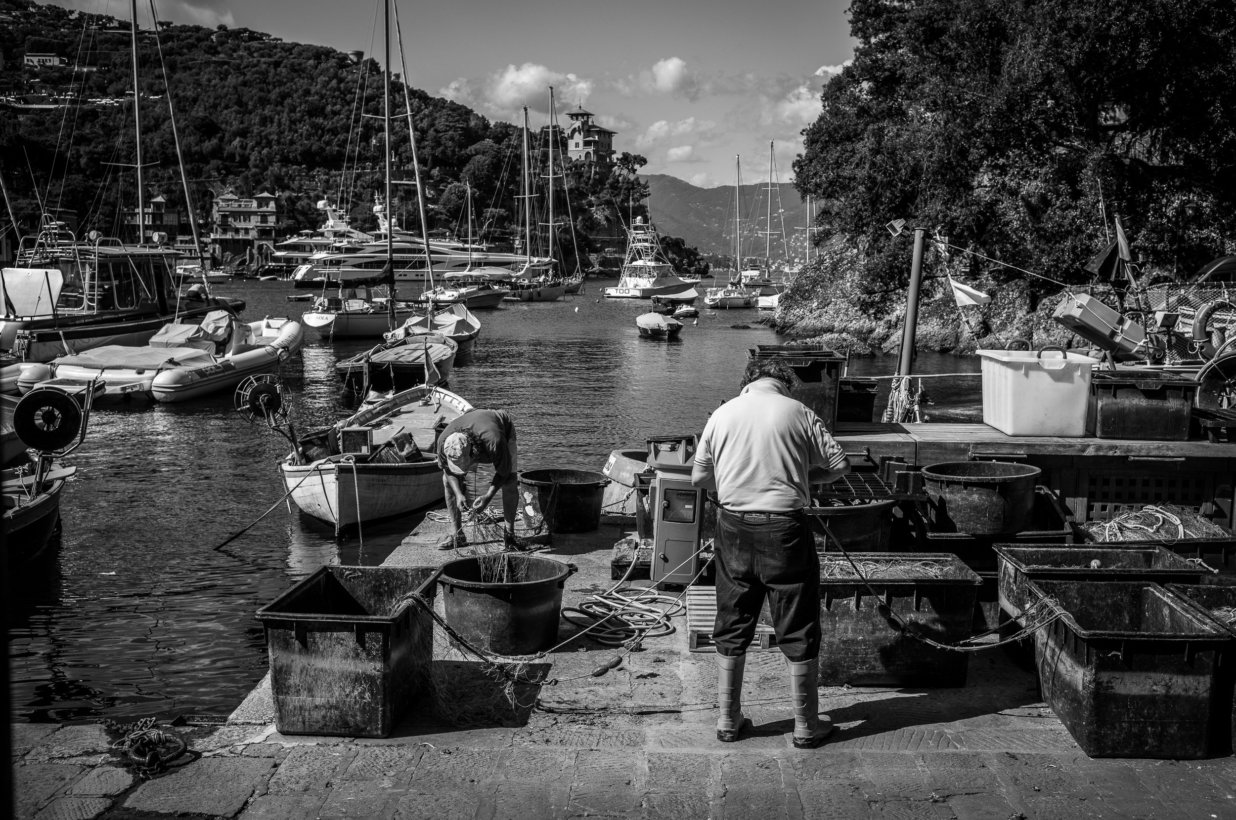Zwei Männer arbeiten an einem Hafen mit mehreren Booten, das Meer ist ruhig, im Hintergrund Berge und Häuser, im Bild ist alles in Schwarzweiß gehalten.
