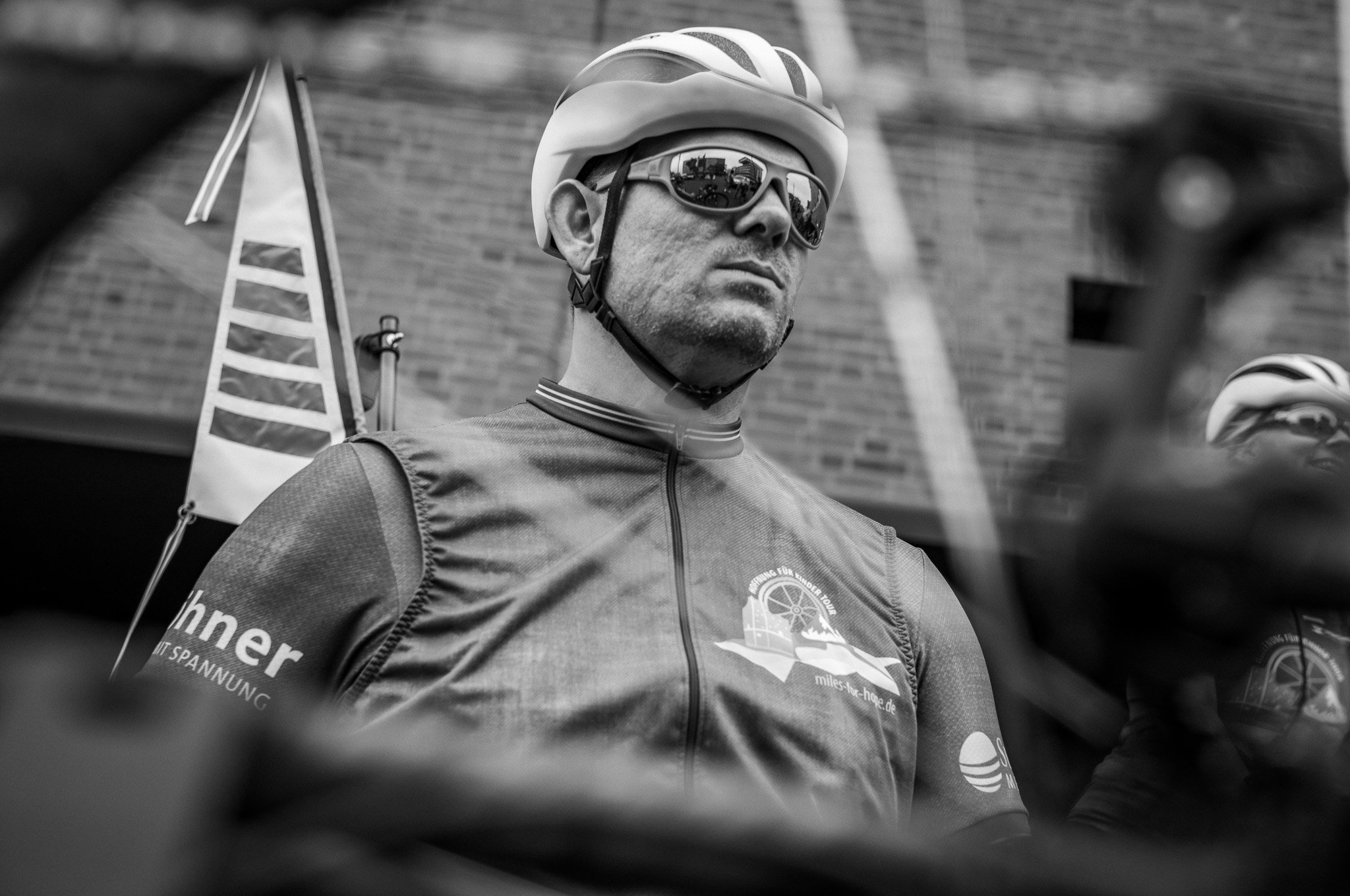 Ein Mann trägt einen Fahrradhelm und Sonnenbrille, trägt eine Radjacke mit einem Logo, im Hintergrund eine Wand und ein Fahrrad. Es ist eine Schwarz-Weiß-Aufnahme, vermutlich bei einer Fahrradveranstaltung.