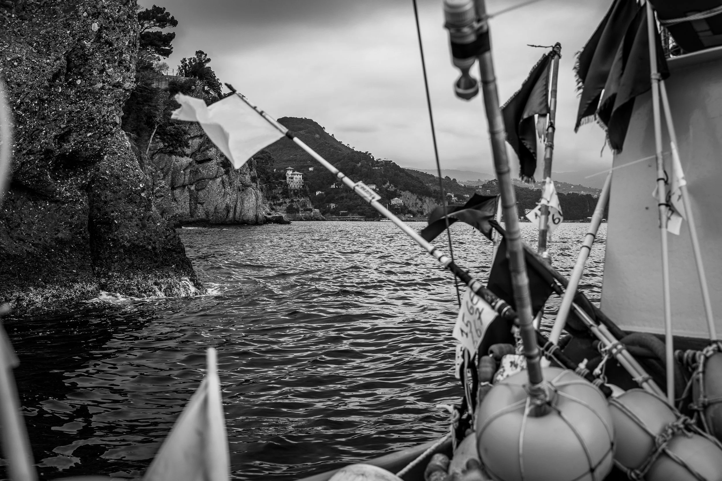 Blick vom Boot auf das Wasser, Felsen an der Küste und Häuser auf den Hügeln im Hintergrund, schwarz-weiß, maritime Szene.