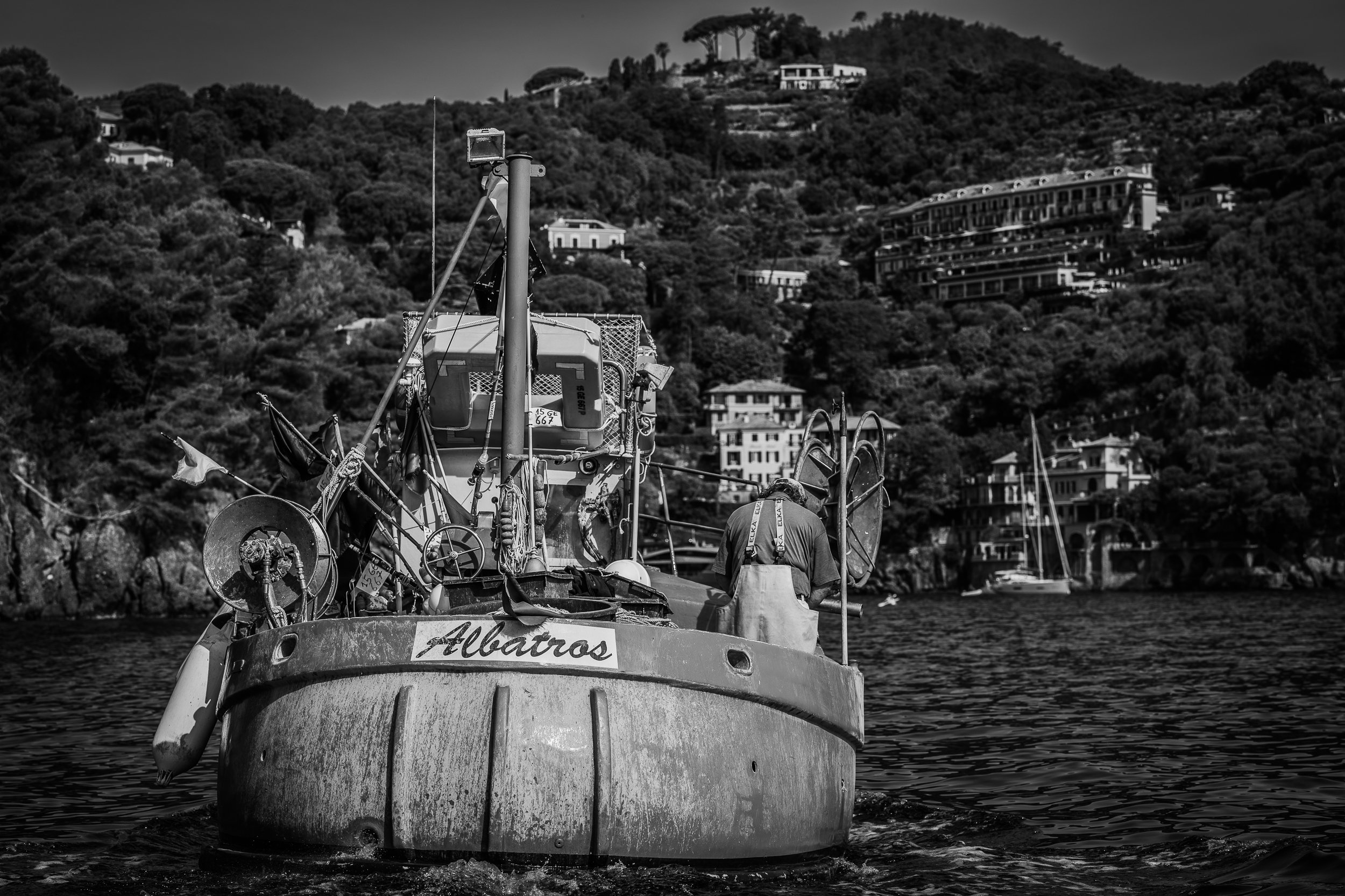 Ein altes Boot namens 'Albatros' auf dem Wasser mit einer Person im Hintergrund, und es gibt Häuser auf einem Hügel im Hintergrund.