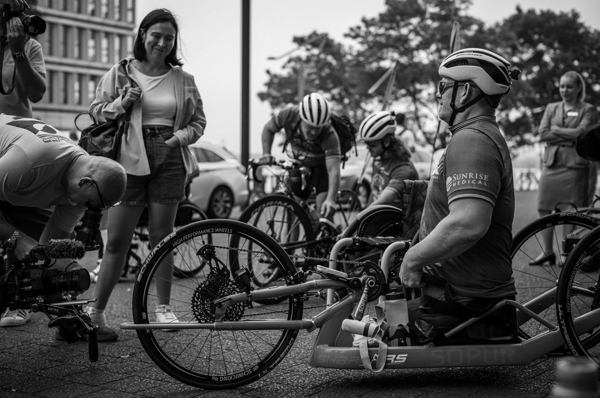 Menschen beim Einschieben eines behindertengerechten Fahrrads auf die Straße, einige tragen Helme, andere schauen zu, im Hintergrund sind Bäume und Autos zu sehen.