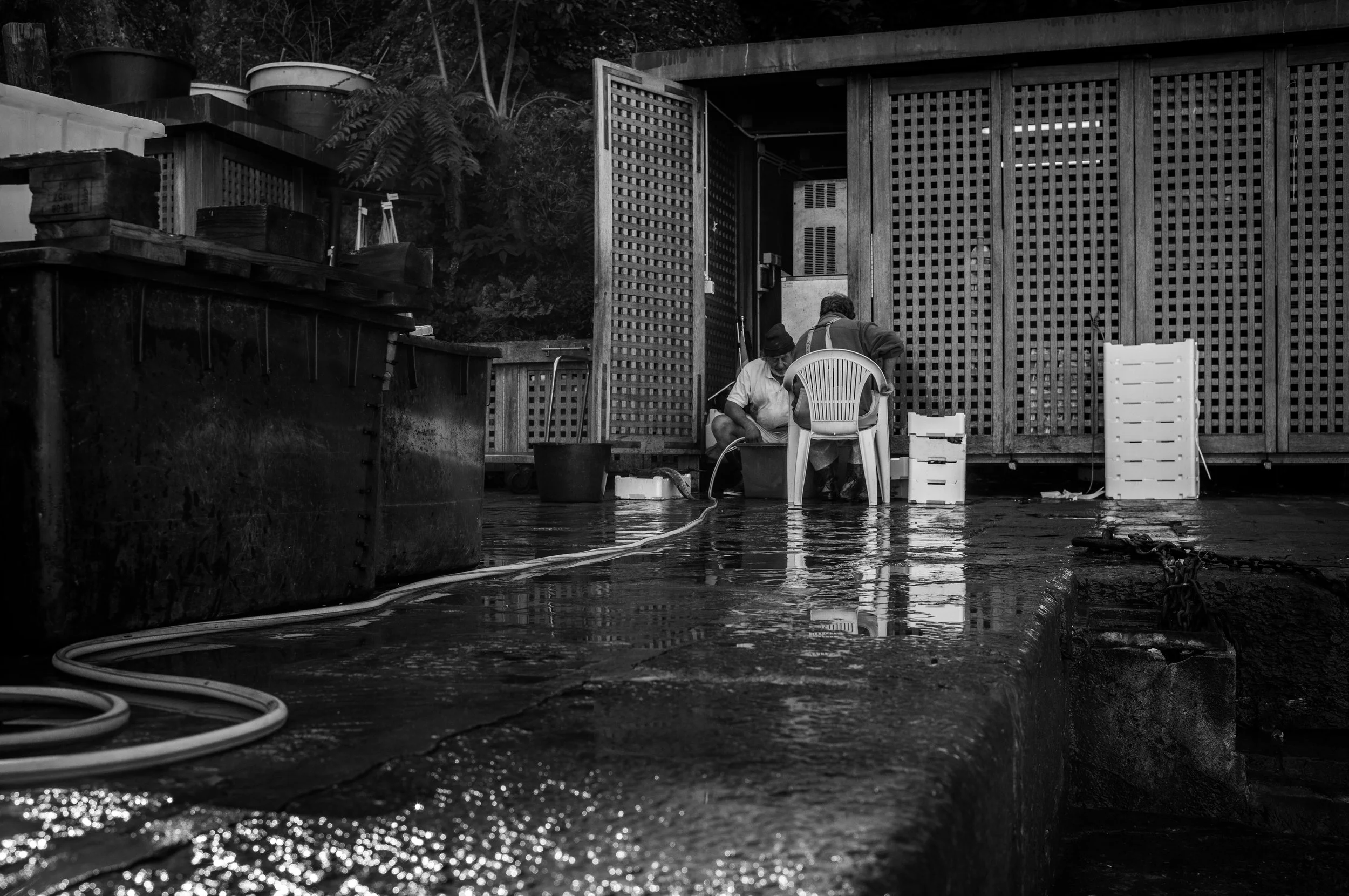 Zwei Menschen arbeiten im Freien an einem Wasseranschluss, einer sitzt in einem Badeschirm, während der andere Wasser an einer Leitung anbringt, mit Wasserpfützen auf dem Boden und Gegenständen im Hintergrund.