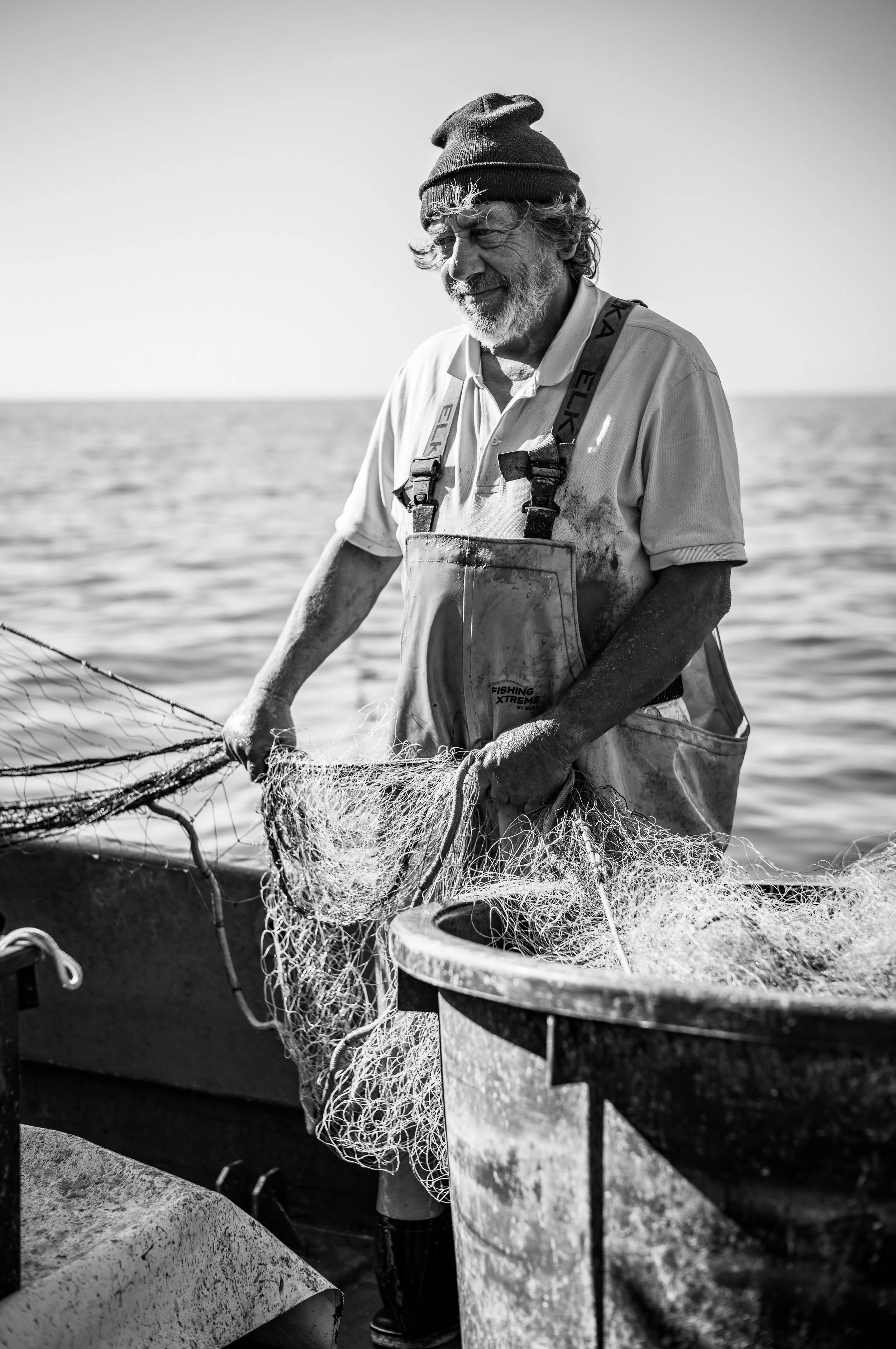 Ein älterer Mann mit weißem Bart und Locken, trägt einen dunklen Hut und eine Arbeitskleidung, während er im Meer einen Fischfang mit einem Netzt macht.