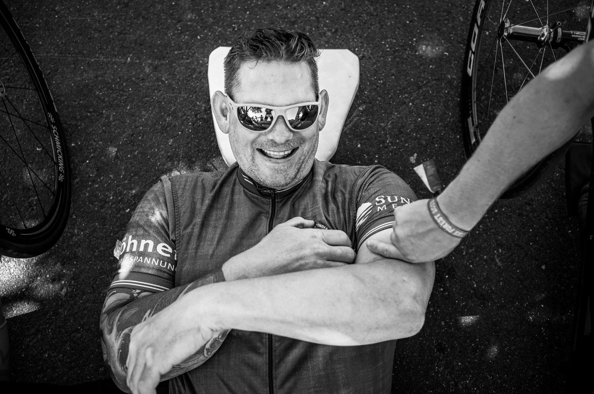 Ein lachender Mann mit Sonnenbrille liegt auf dem Rücken, während jemand seinen Arm in die Nähe seiner Schulter drückt. Er trägt einen Radtrikot und befindet sich neben Fahrradfelgen auf einer dunklen Oberfläche.