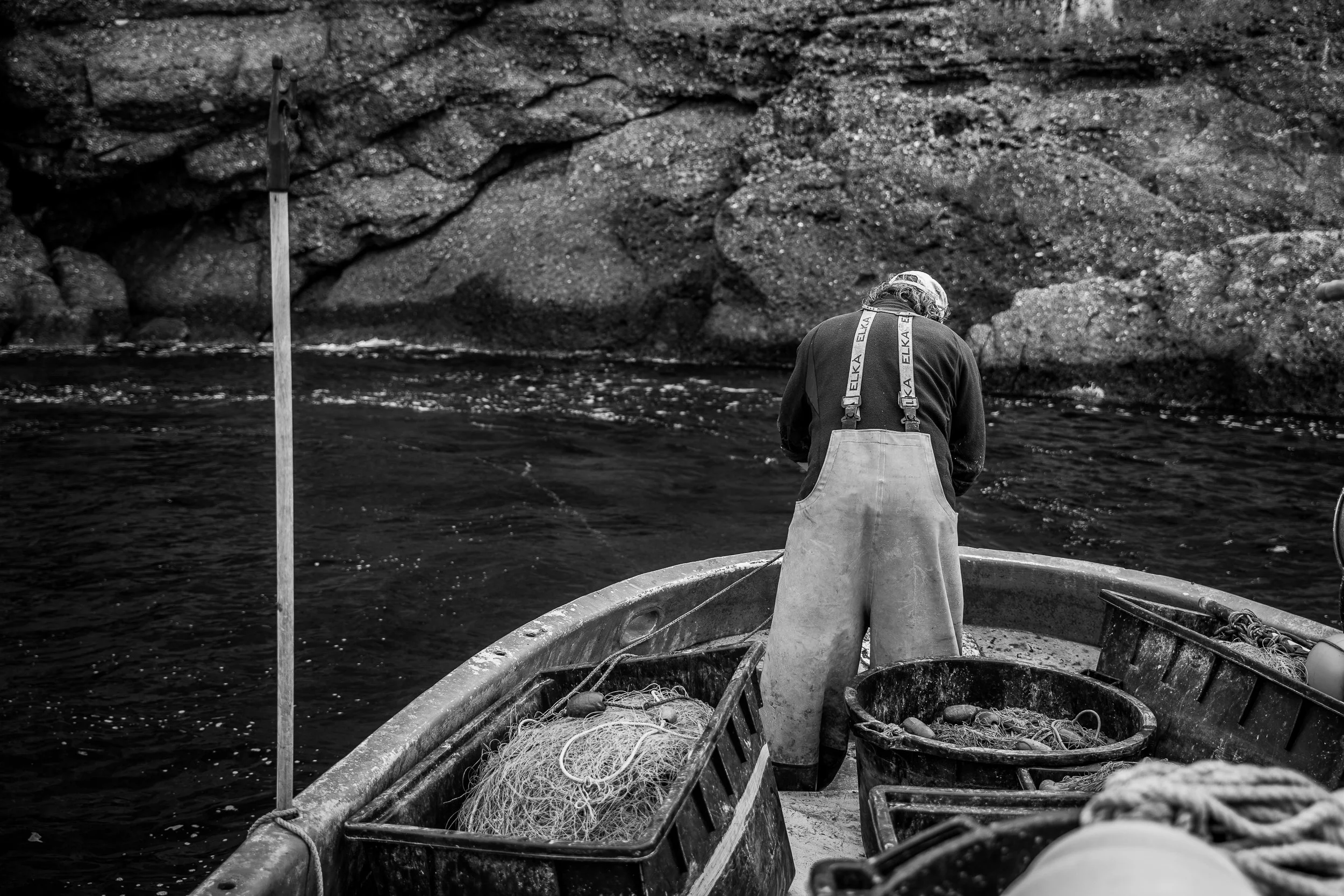 Ein Fischer in overalls steht in einem Boot auf einem Wasser, umgeben von Netzen und Ausrüstung, vor felsiger Küste, aufgenommen in Schwarz-Weiß.