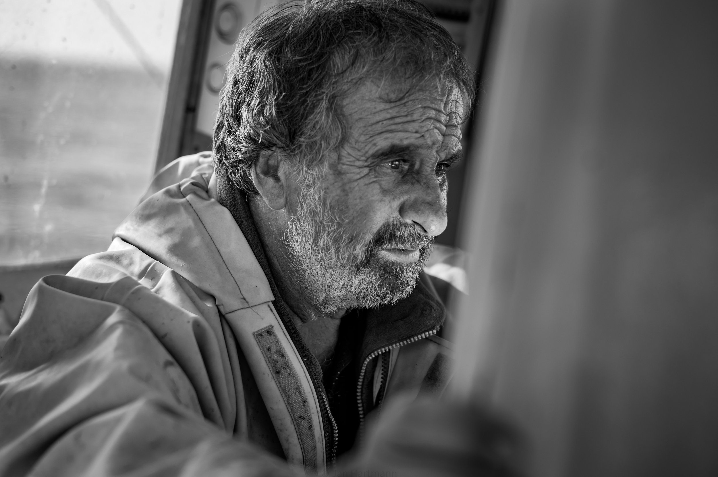 Ein älterer Mann mit grauem Haar und Bart schaut nach rechts, trägt eine Jacke, im Hintergrund eine Fensterfront.
