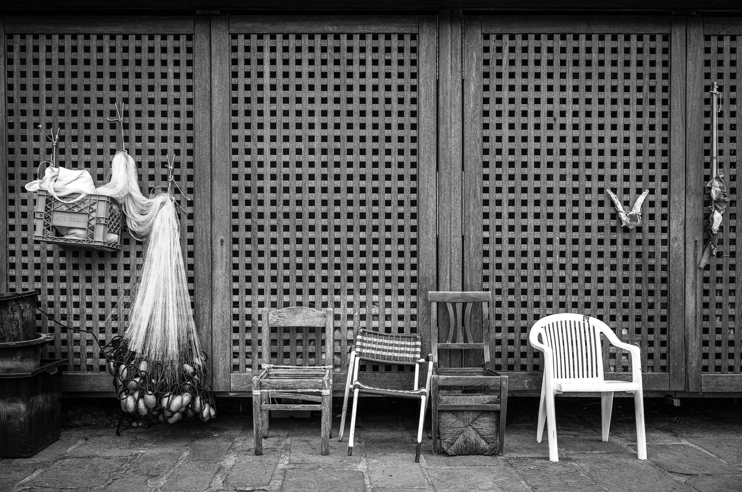 Fünf verschiedene Stühle gegen eine Holzwand mit Gittermuster, darunter hängen Trend-Accessoires, inklusive Pelz- und Plastikboxen, eine Vogelskulptur und eine Wasserpumpe.