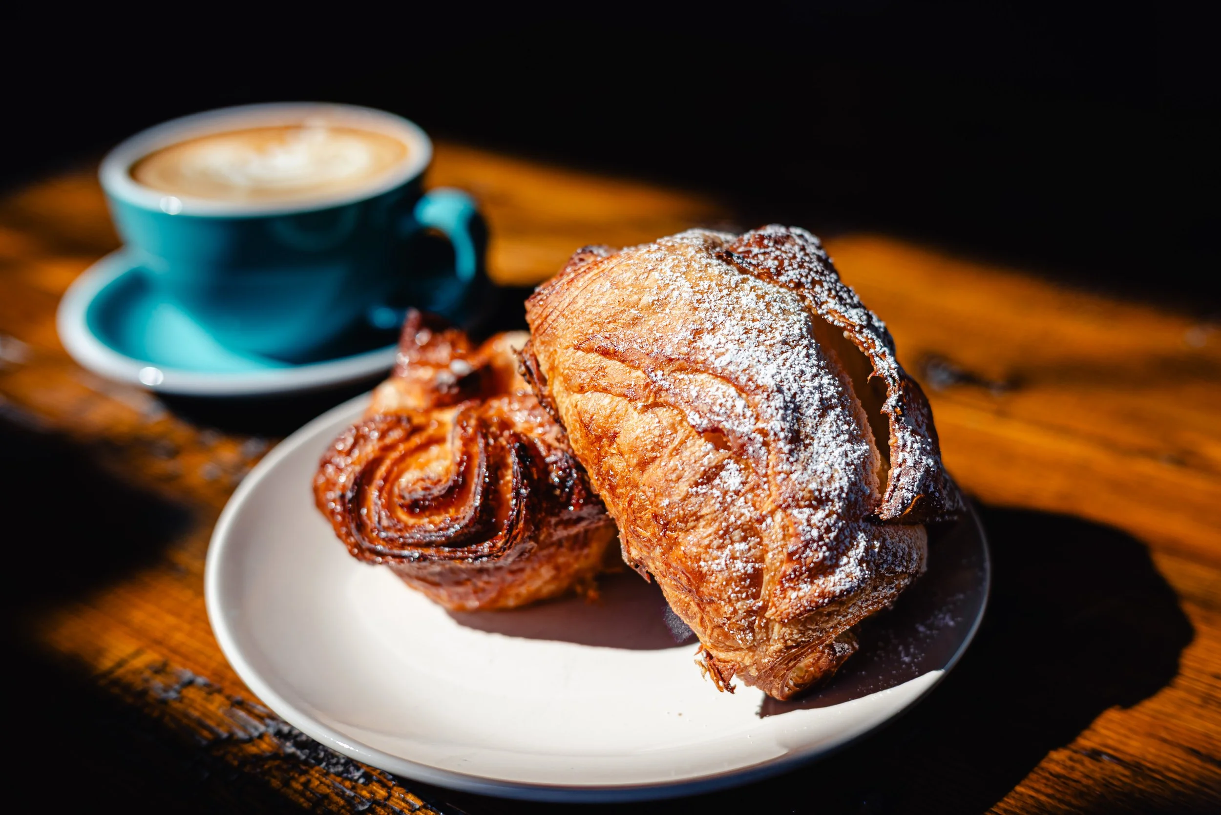 A white plate holds a croissant dusted with powdered sugar and a cinnamon roll. In the background, a blue cup of coffee with foam sits on a matching saucer on a wooden table.