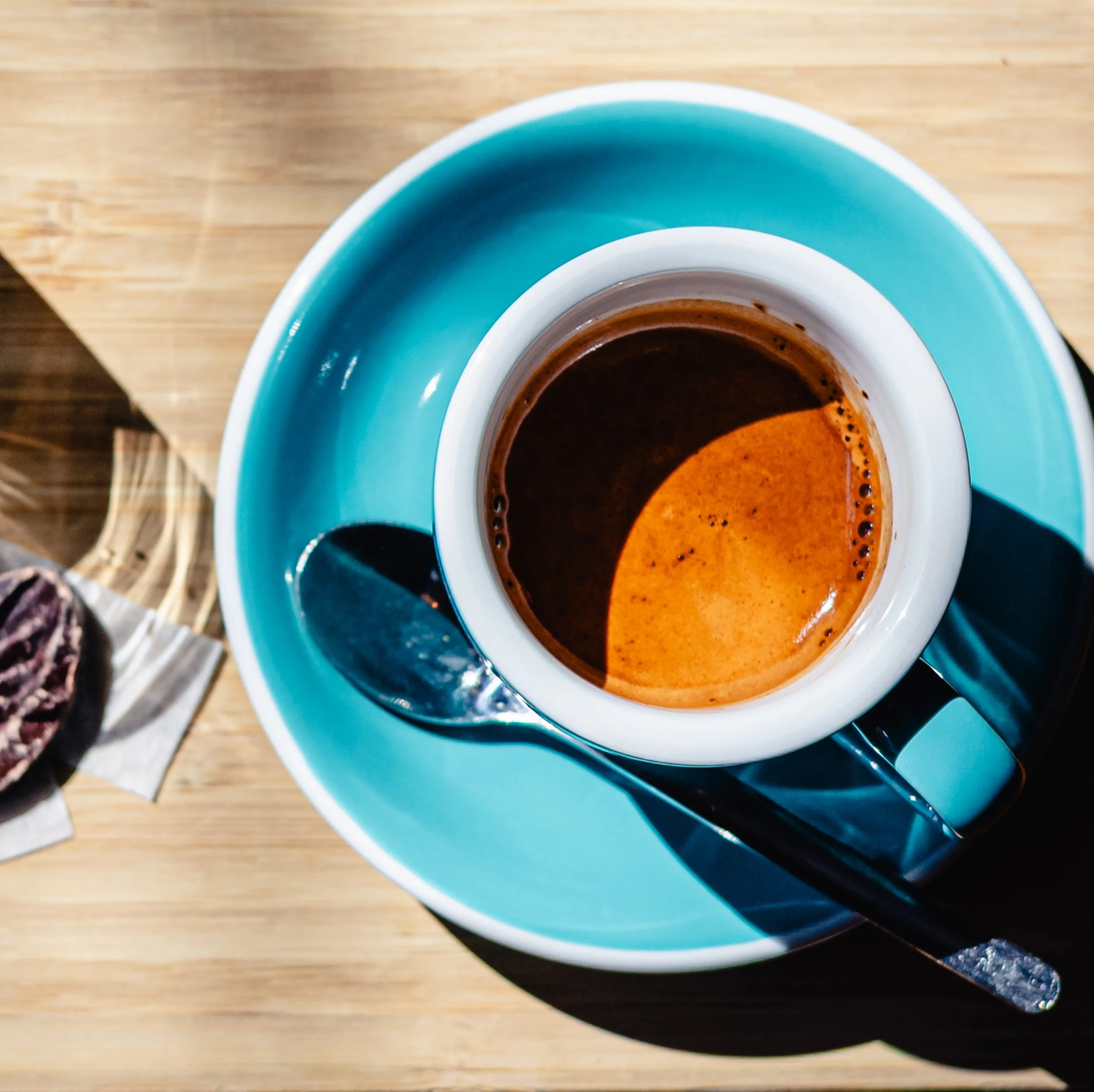 A cup of black coffee on a teal saucer with a metal spoon, on a wooden table.