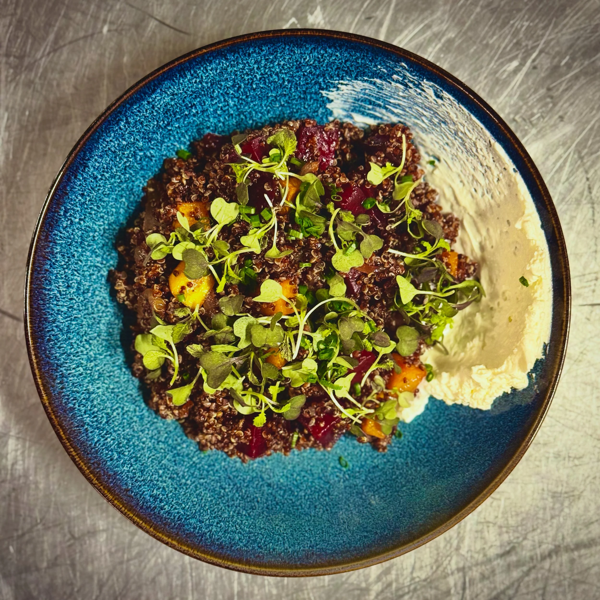 A bowl of cooked quinoa with diced vegetables and microgreens on top, placed on a gray surface.