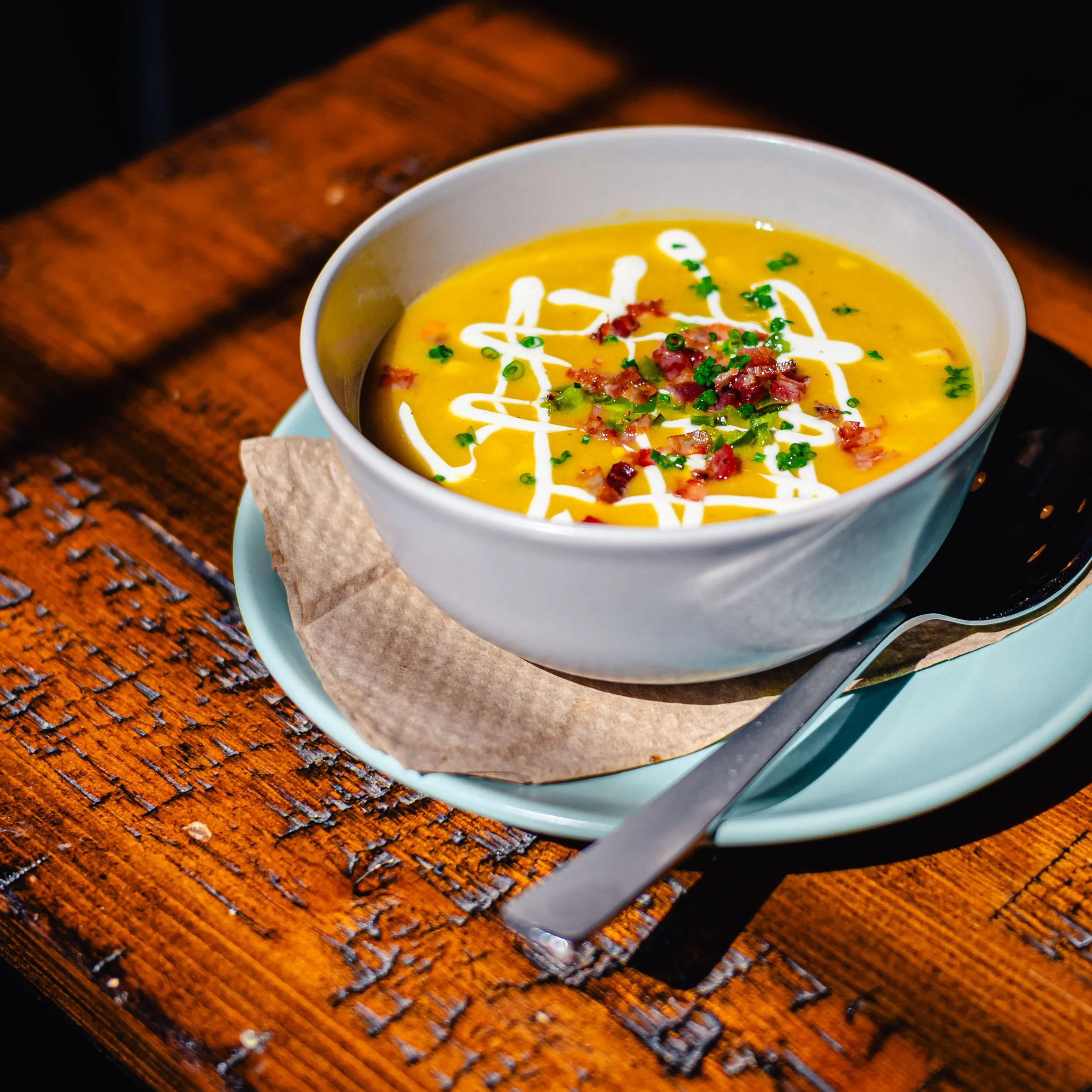 Bowl of creamy yellow soup garnished with bacon bits, chopped green onions, and drizzled with sour cream, served on a light blue plate with a metal spoon on a wooden table.