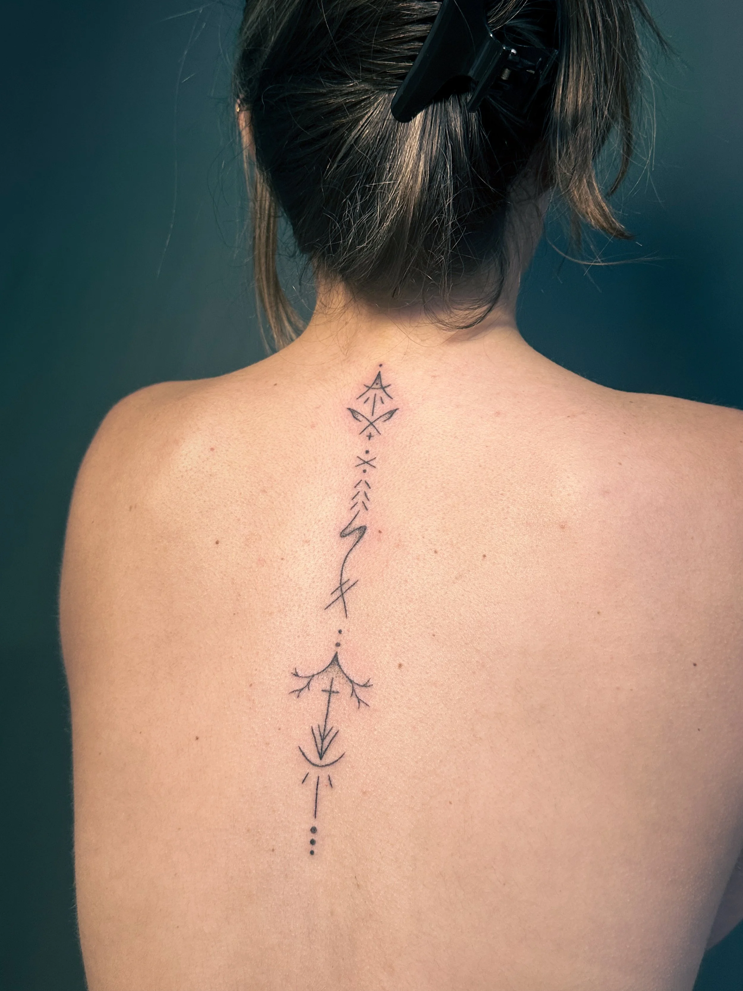 Back of a person with brown hair, wearing a black hair clip, and a line tattoo running down the spine with geometric and arrow-like elements.