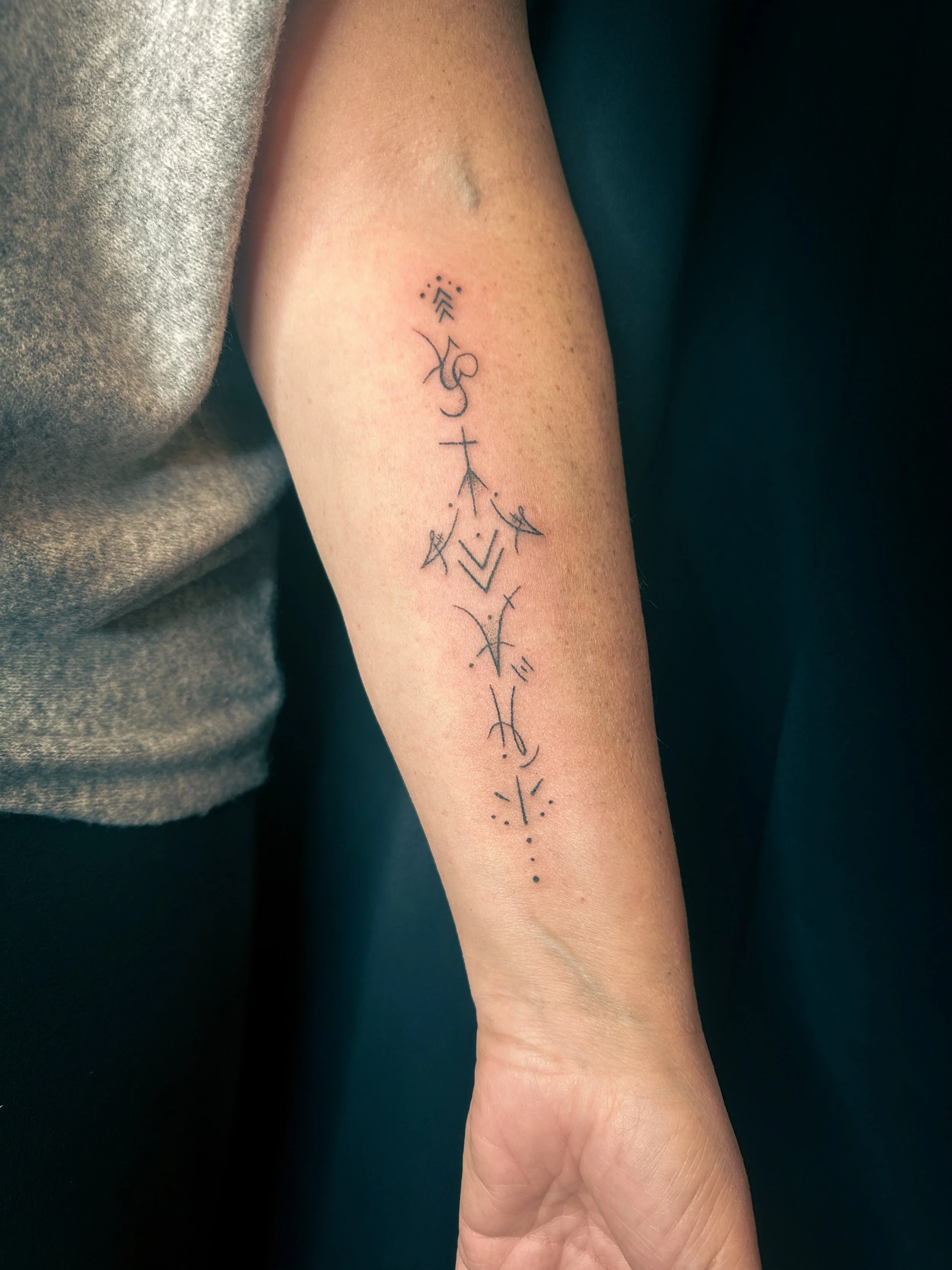 A person's forearm with a modern, minimalistic black tattoo featuring geometric shapes and lines, some dotted and arrow-like elements.